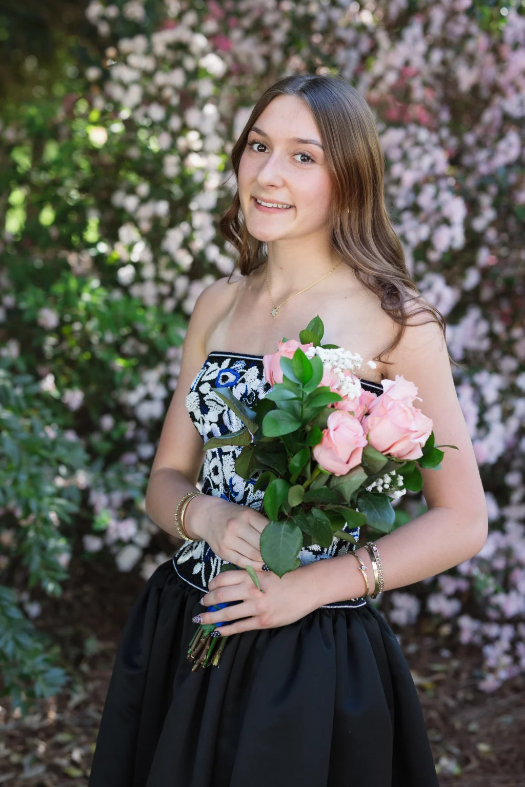 BJA Jr/Sr Banquet Prom at Sue Simpson Park: Young woman in a strapless black and white dress holding a bouquet of pink roses, standing outdoors in front of blooming bushes.