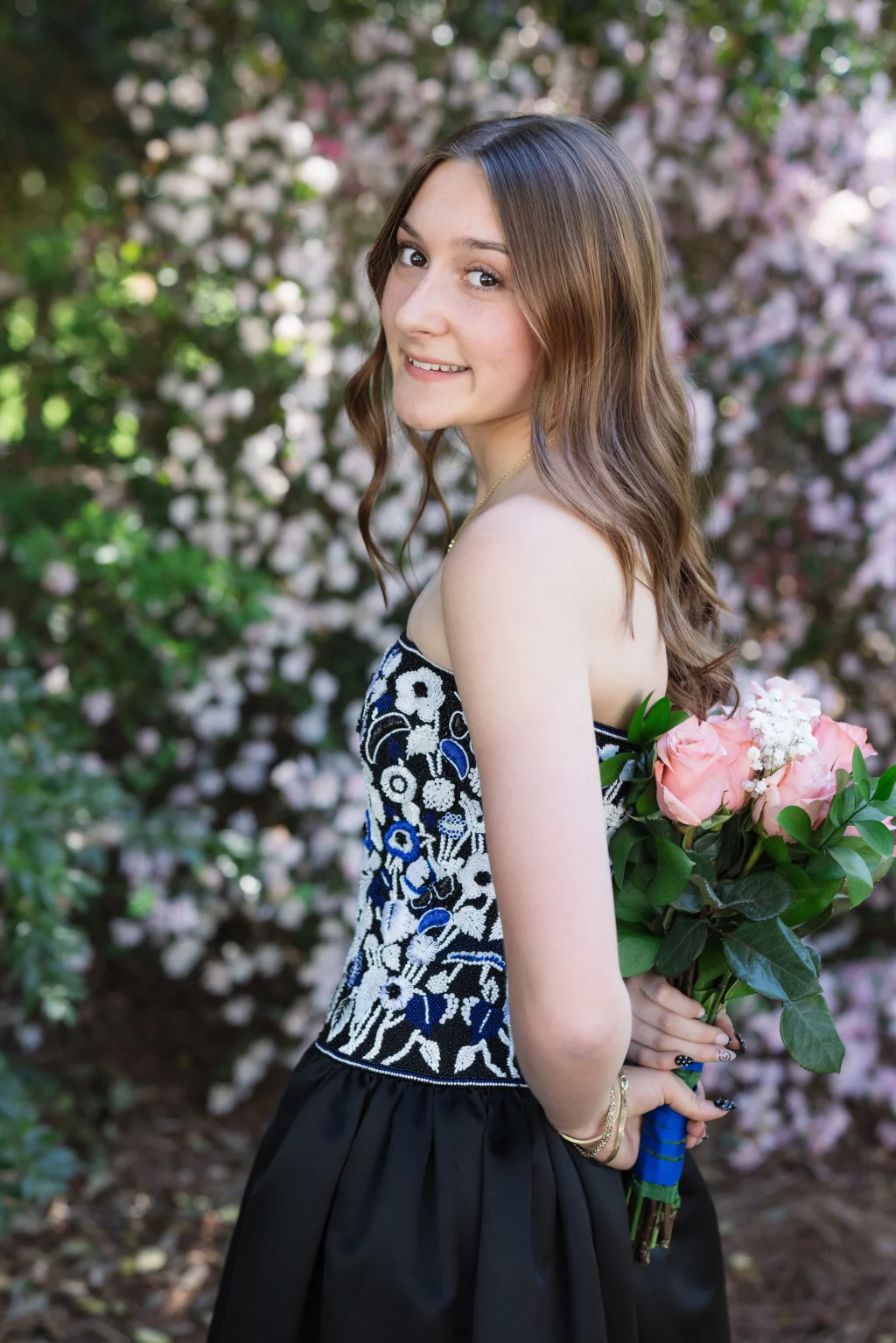 BJA Jr/Sr Banquet Prom at Sue Simpson Park: Young woman with wavy brown hair wearing a strapless floral-patterned dress, holding a bouquet of pink roses behind her back, standing outdoors in front of blooming bushes.
