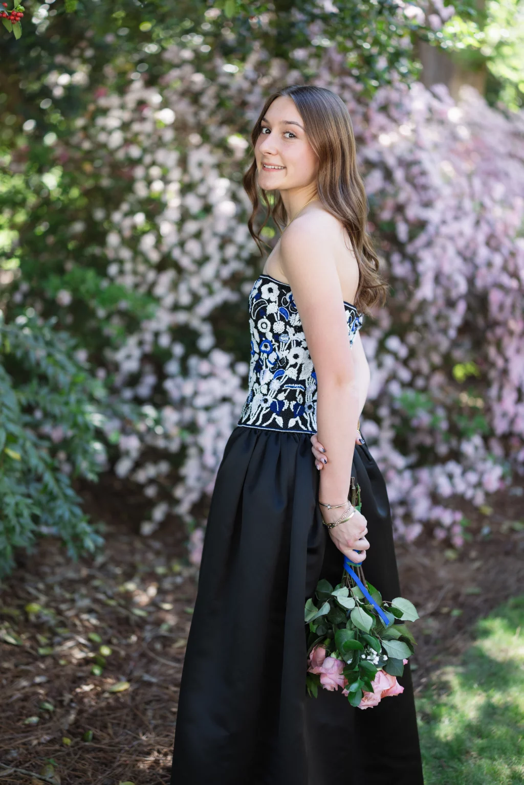 BJA Jr/Sr Banquet Prom at Sue Simpson Park: Young woman in a formal black and floral dress standing outdoors, holding a bouquet of pink roses with a blue ribbon, smiling with blooming pink flowers in the background.