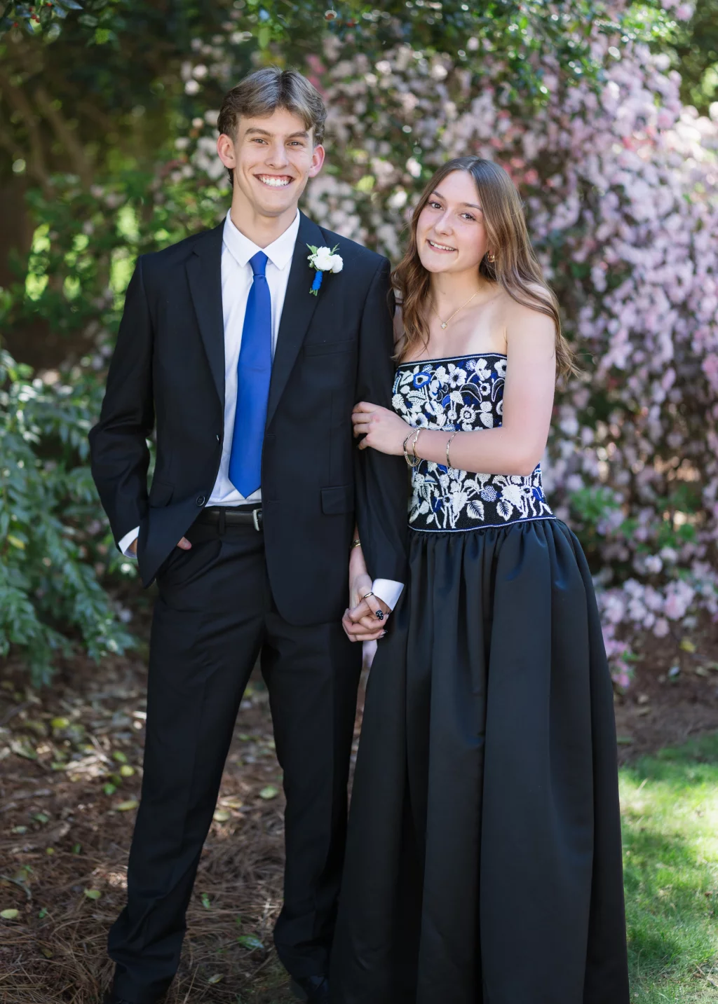 BJA Jr/Sr Banquet Prom at Sue Simpson Park: A young man in a black suit with a blue tie and a boutonniere stands next to a young woman in a black strapless dress with white floral embroidery. They are smiling, holding hands, and standing outdoors in front of blooming pink flowers and greenery.