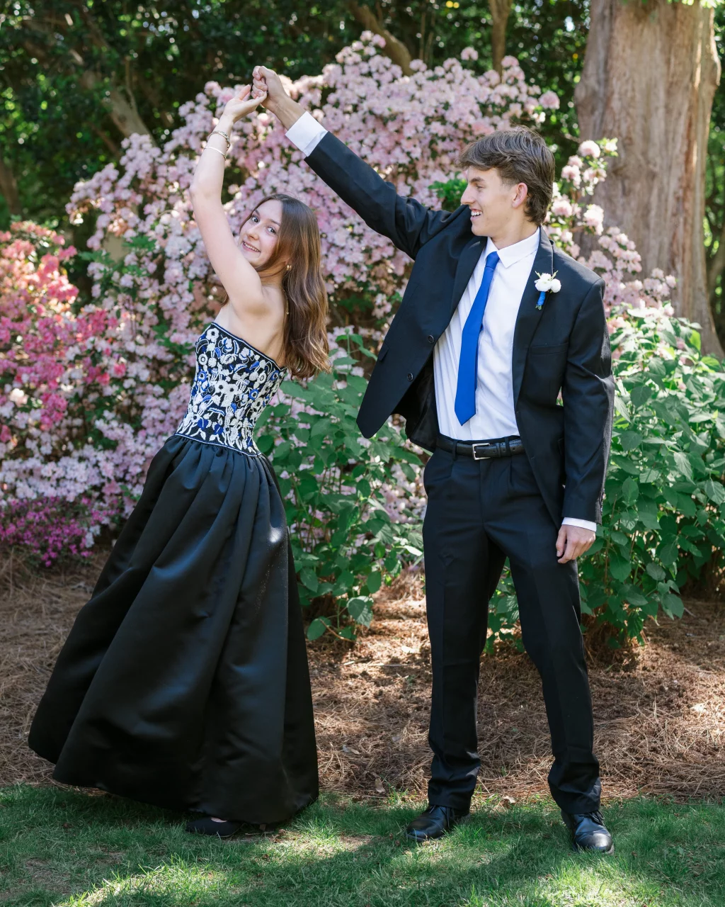 BJA Jr/Sr Banquet Prom at Sue Simpson Park: A young woman in a strapless black and white gown and a young man in a black suit with a blue tie dance together outside, surrounded by blooming pink flowers and greenery.