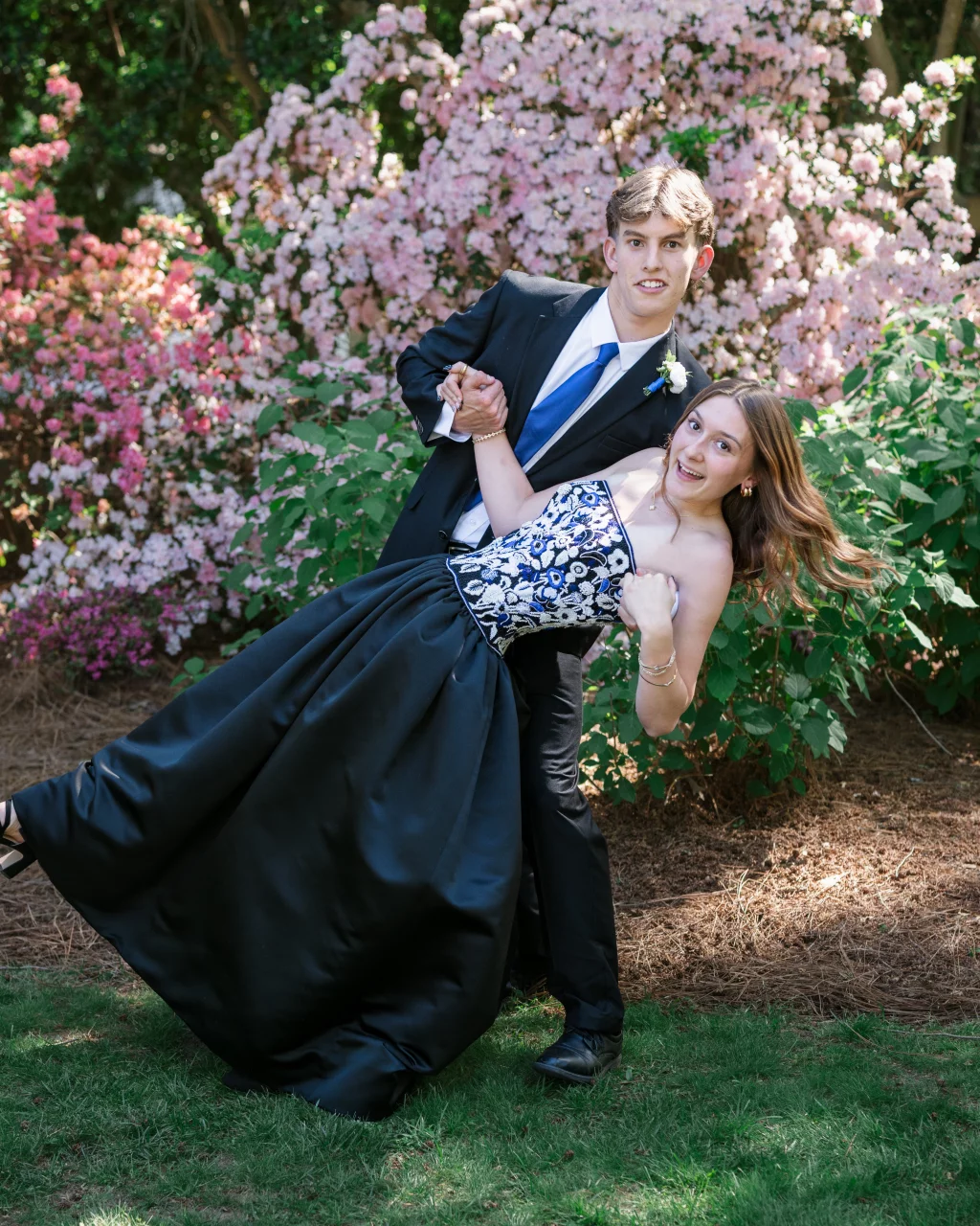 BJA Jr/Sr Banquet Prom at Sue Simpson Park: A young couple dressed for a formal event poses outdoors; the young man in a suit and blue tie dips the young woman, who wears a strapless black and white gown, in front of blooming pink bushes.