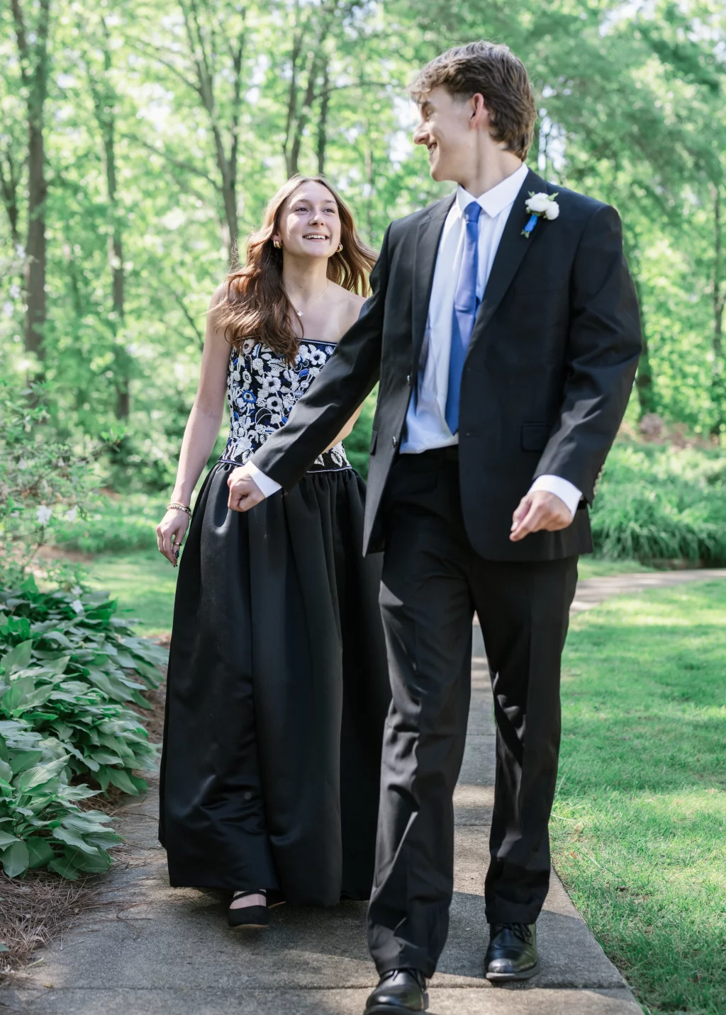 BJA Jr/Sr Banquet Prom at Sue Simpson Park: A young couple dressed in formal attire walks hand-in-hand on a garden path, smiling at each other. The background features lush green trees and plants.