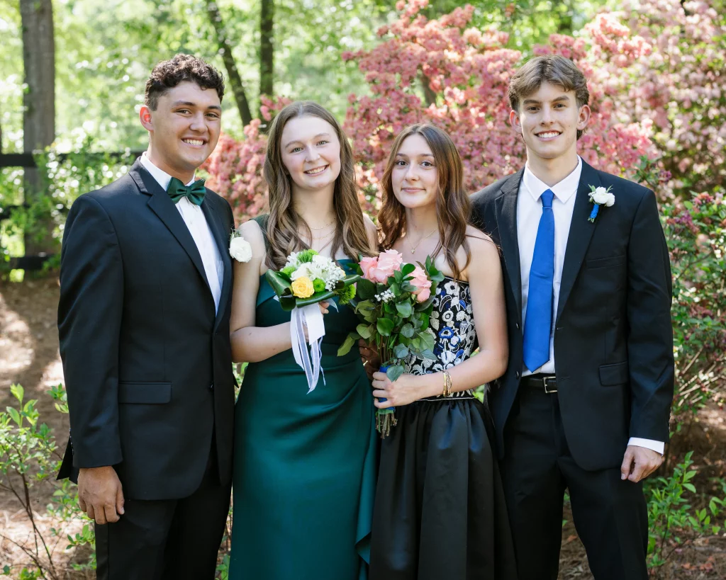 BJA Jr/Sr Banquet Prom at Sue Simpson Park: Four teenagers dressed in formal attire stand together outdoors, smiling, with two holding flower bouquets. Pink flowering bushes and greenery are in the background.