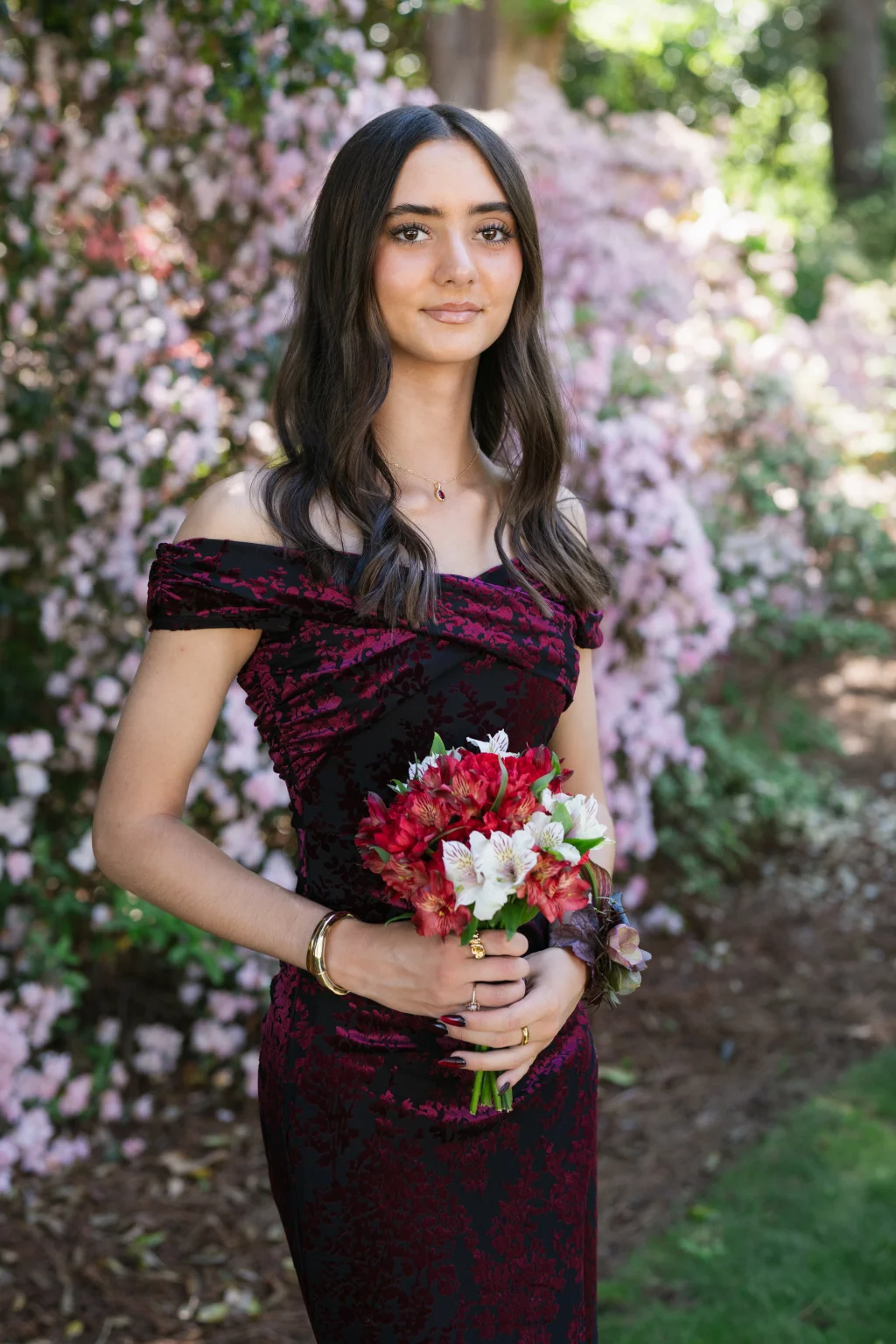 BJA Jr/Sr Banquet Prom at Sue Simpson Park: Young woman in a dark red and black off-the-shoulder dress holding a bouquet of red and white flowers, standing outdoors in front of blooming pink bushes.