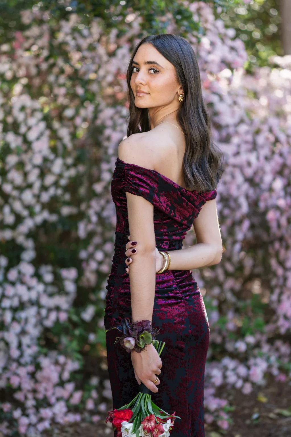 BJA Jr/Sr Banquet Prom at Sue Simpson Park: Young woman in an off-the-shoulder burgundy floral dress holding a bouquet of red and white flowers, standing in front of blooming pink bushes.
