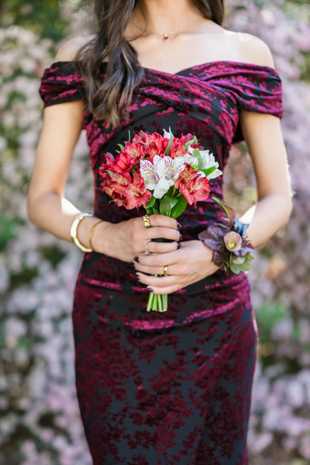 BJA Jr/Sr Banquet Prom at Sue Simpson Park: Woman in an off-the-shoulder, dark red and black dress holding a bouquet of red and white flowers, with a floral corsage on her wrist, standing in front of a blurred floral background.
