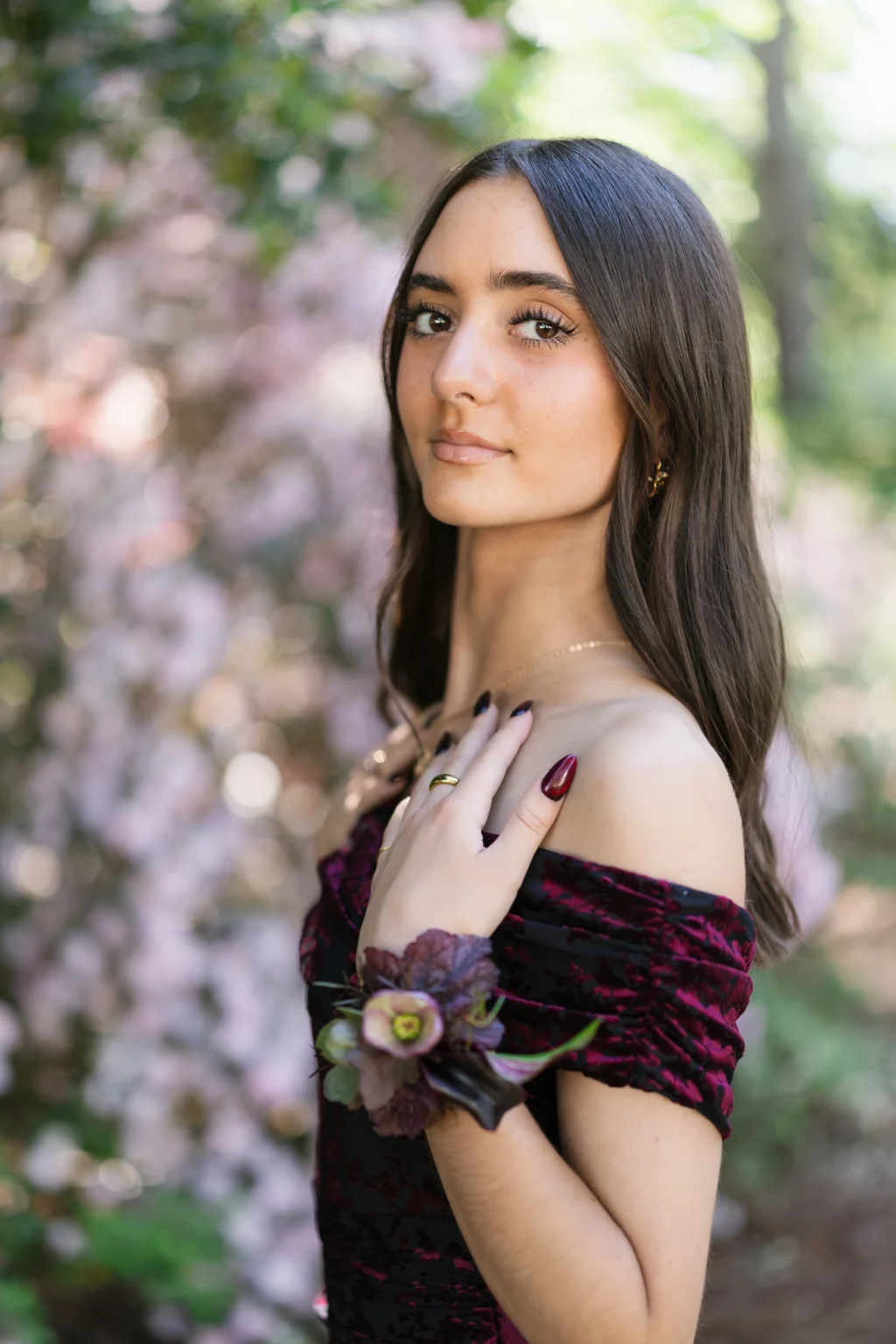 BJA Jr/Sr Banquet Prom at Sue Simpson Park: Young woman with long brown hair wearing an off-shoulder burgundy dress and a purple floral corsage, standing outdoors in front of blurred pink flowers and greenery.