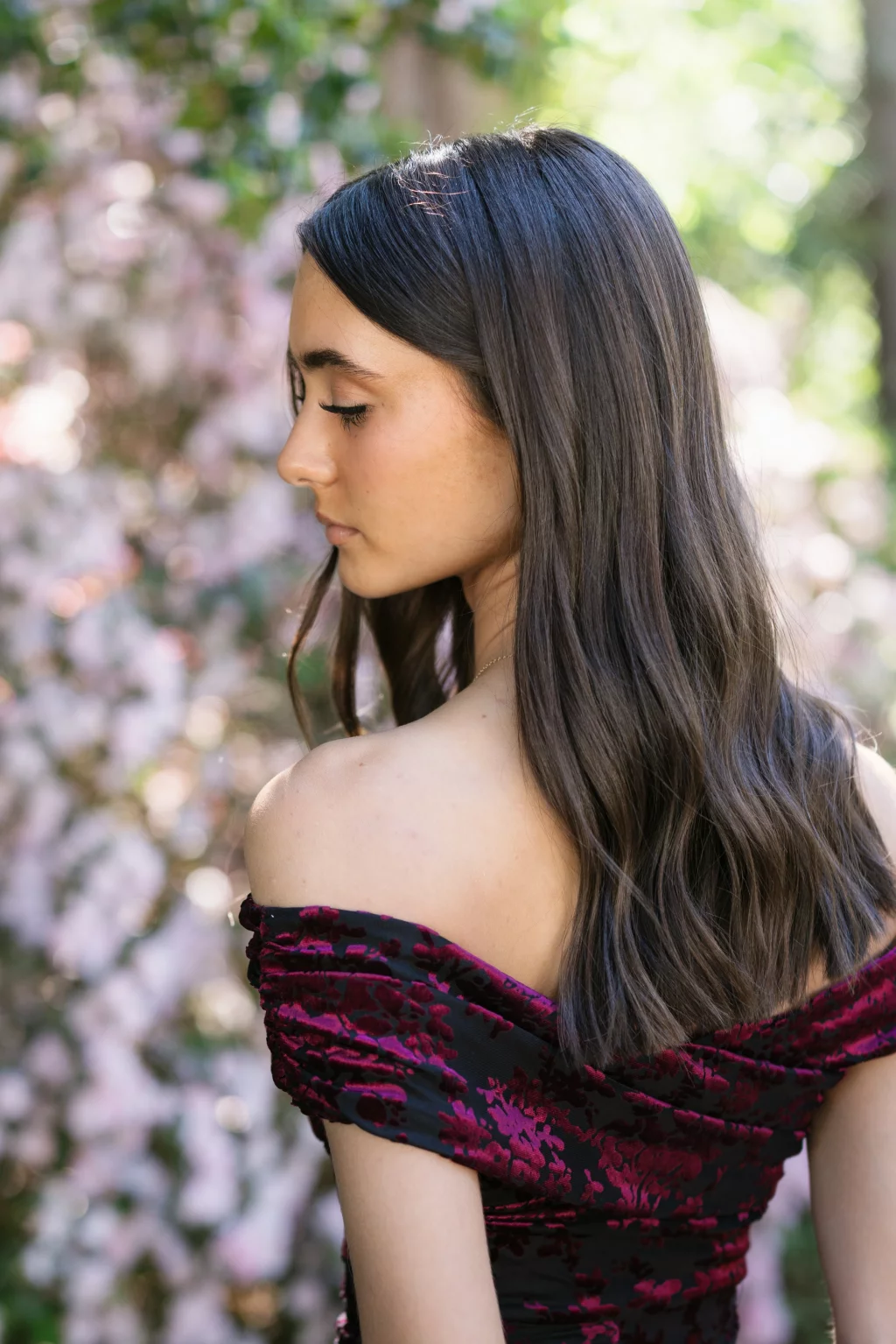 BJA Jr/Sr Banquet Prom at Sue Simpson Park: A young woman with long brown hair, wearing an off-the-shoulder dress with a dark red floral pattern, stands outdoors in front of blurred pink and green foliage, looking down and to the side.