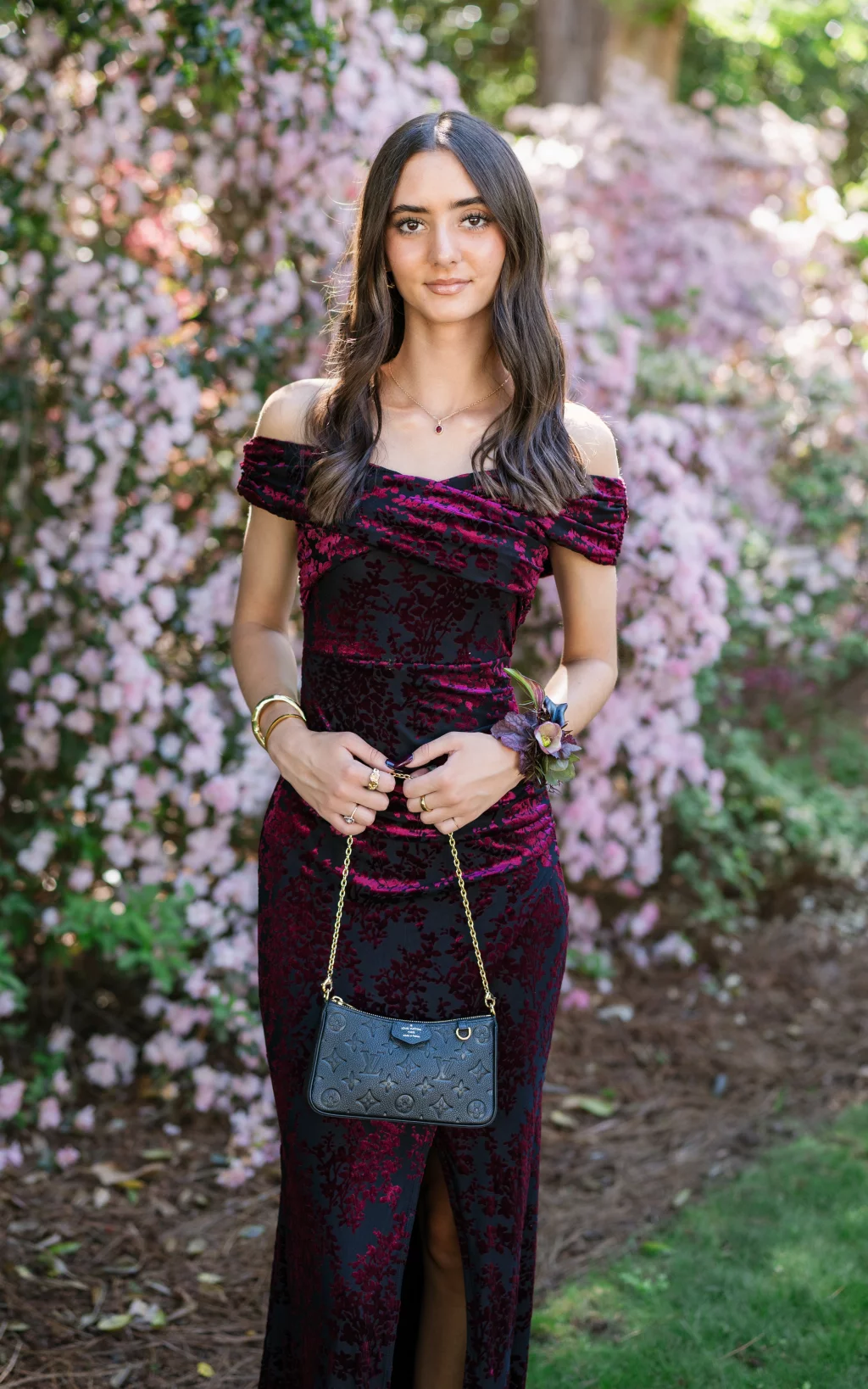 BJA Jr/Sr Banquet Prom at Sue Simpson Park: Young woman standing outdoors in front of blooming pink flowers, wearing a dark red and black off-the-shoulder dress, holding a small black purse with a gold chain, and wearing a floral corsage on her wrist.