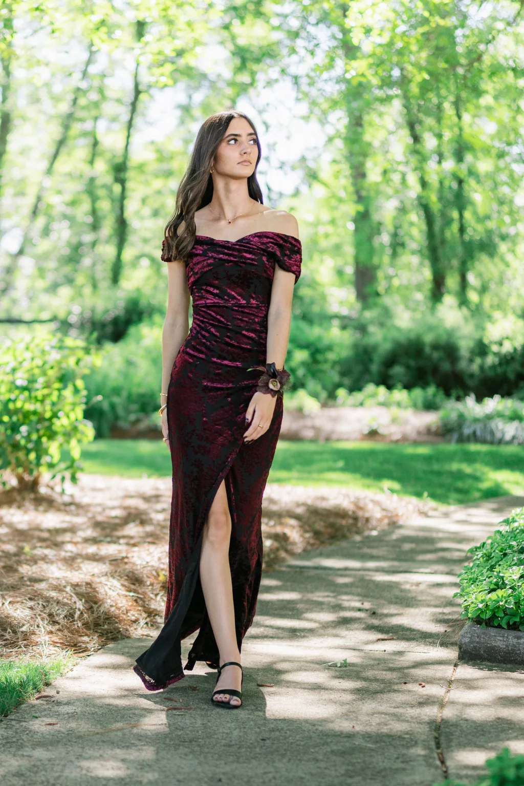 BJA Jr/Sr Banquet Prom at Sue Simpson Park: Young woman in a burgundy off-the-shoulder gown with a corsage, walking on a shaded garden path surrounded by greenery.