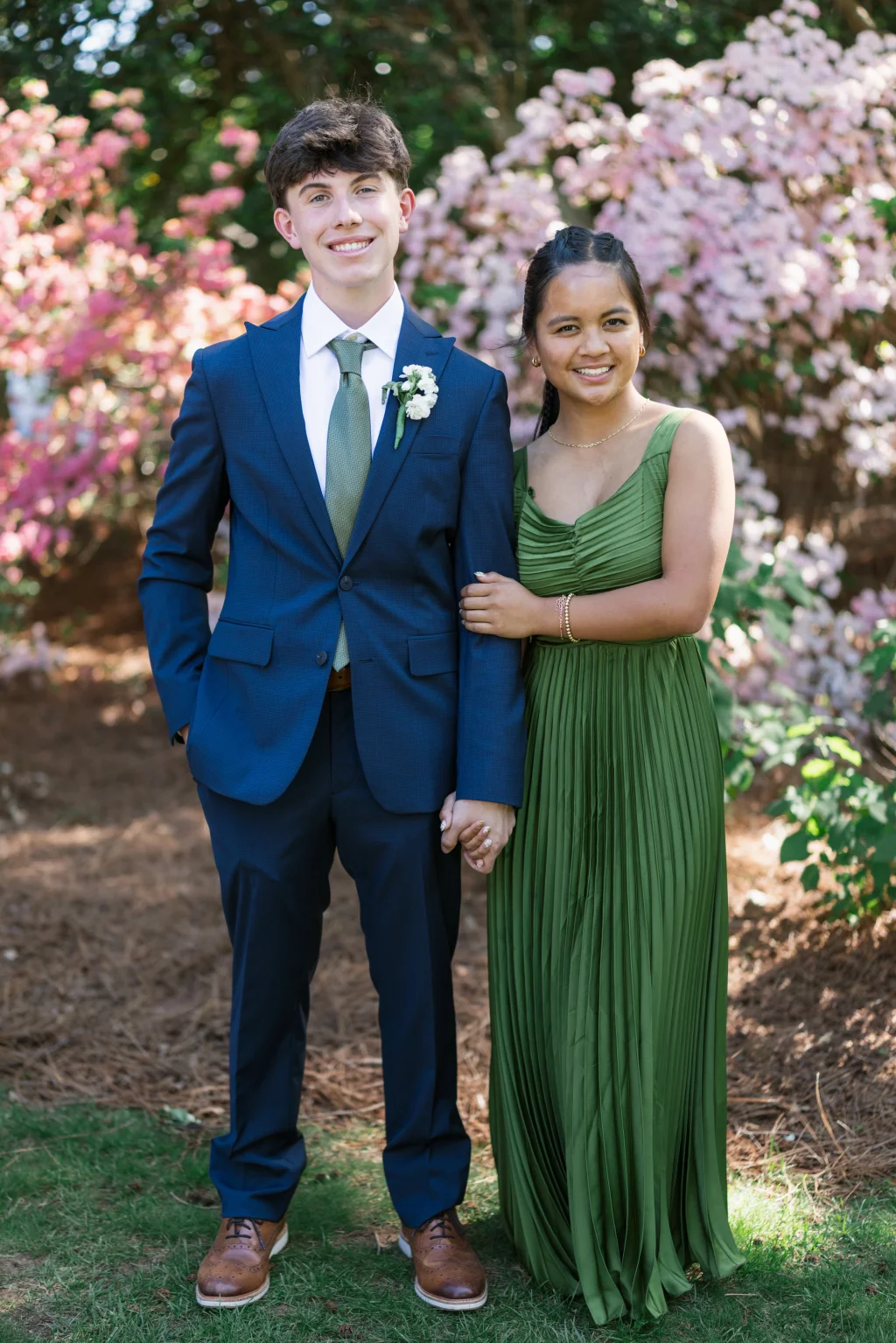 BJA Jr/Sr Banquet Prom at Sue Simpson Park: Two teenagers dressed formally, standing together outdoors in front of blooming pink bushes. The boy wears a dark blue suit, white shirt, green tie, and brown shoes, with a boutonni&egrave;re on his lapel. The girl wears a long, pleated green dress and holds the boy&rsquo;s arm, both smiling at the camera.