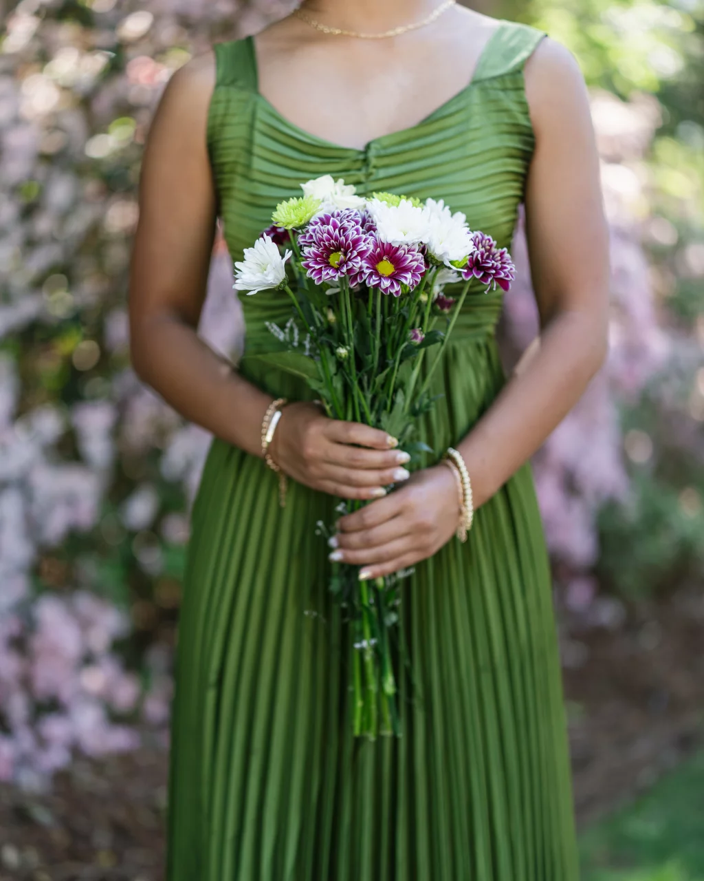 BJA Jr/Sr Banquet Prom at Sue Simpson Park: A person in a green pleated dress holding a bouquet of white, purple, and yellow flowers, standing outdoors with blurred pink flowers in the background.