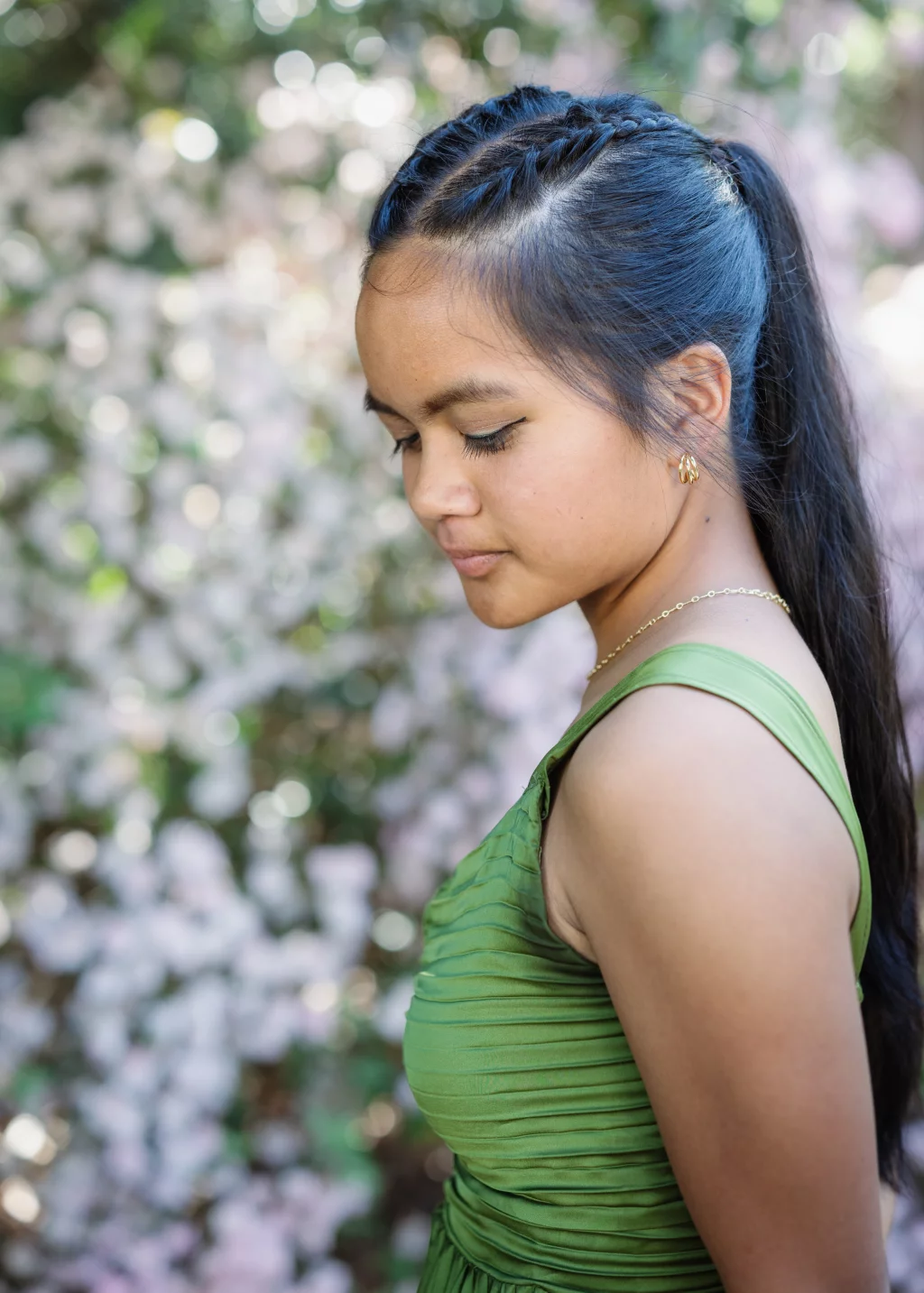 BJA Jr/Sr Banquet Prom at Sue Simpson Park: A young woman in a green dress stands in profile, looking down, with long dark hair styled in braids and a ponytail. She is outdoors, with a blurred background of light pink and white flowers.