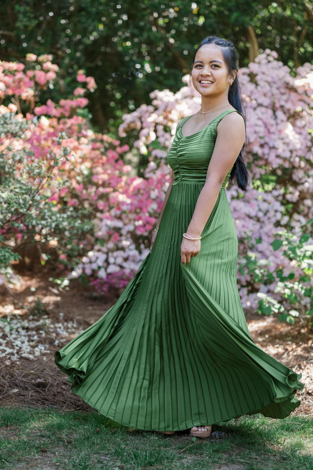 BJA Jr/Sr Banquet Prom at Sue Simpson Park: A young woman in a flowing, pleated green dress stands outdoors, smiling and twirling, surrounded by blooming pink and white flowers and green foliage.