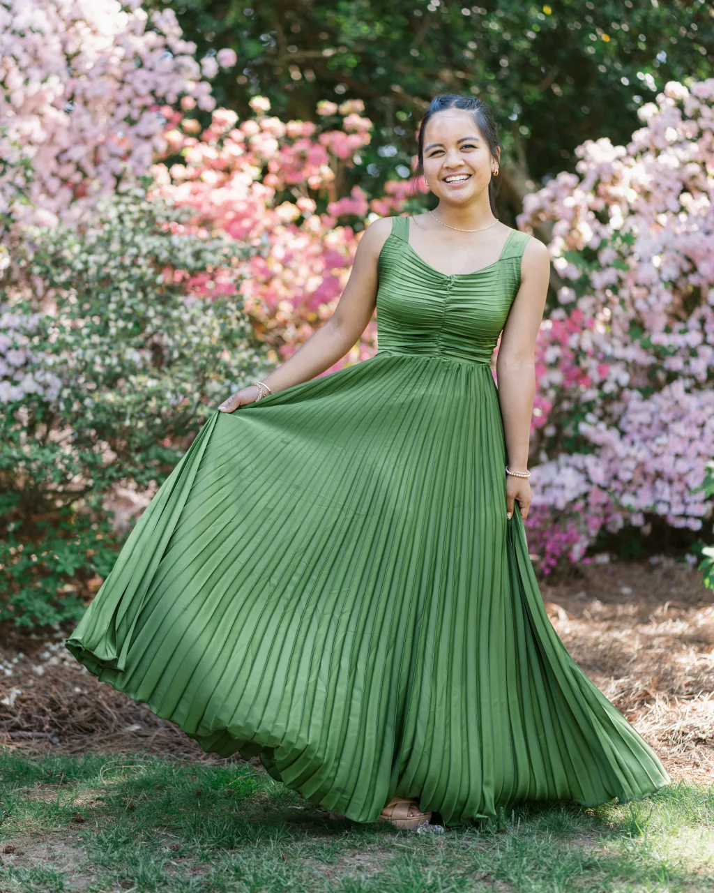 BJA Jr/Sr Banquet Prom at Sue Simpson Park: Smiling young woman in a flowing green pleated dress stands outdoors, holding out her skirt, with blooming pink and white flowers in the background.