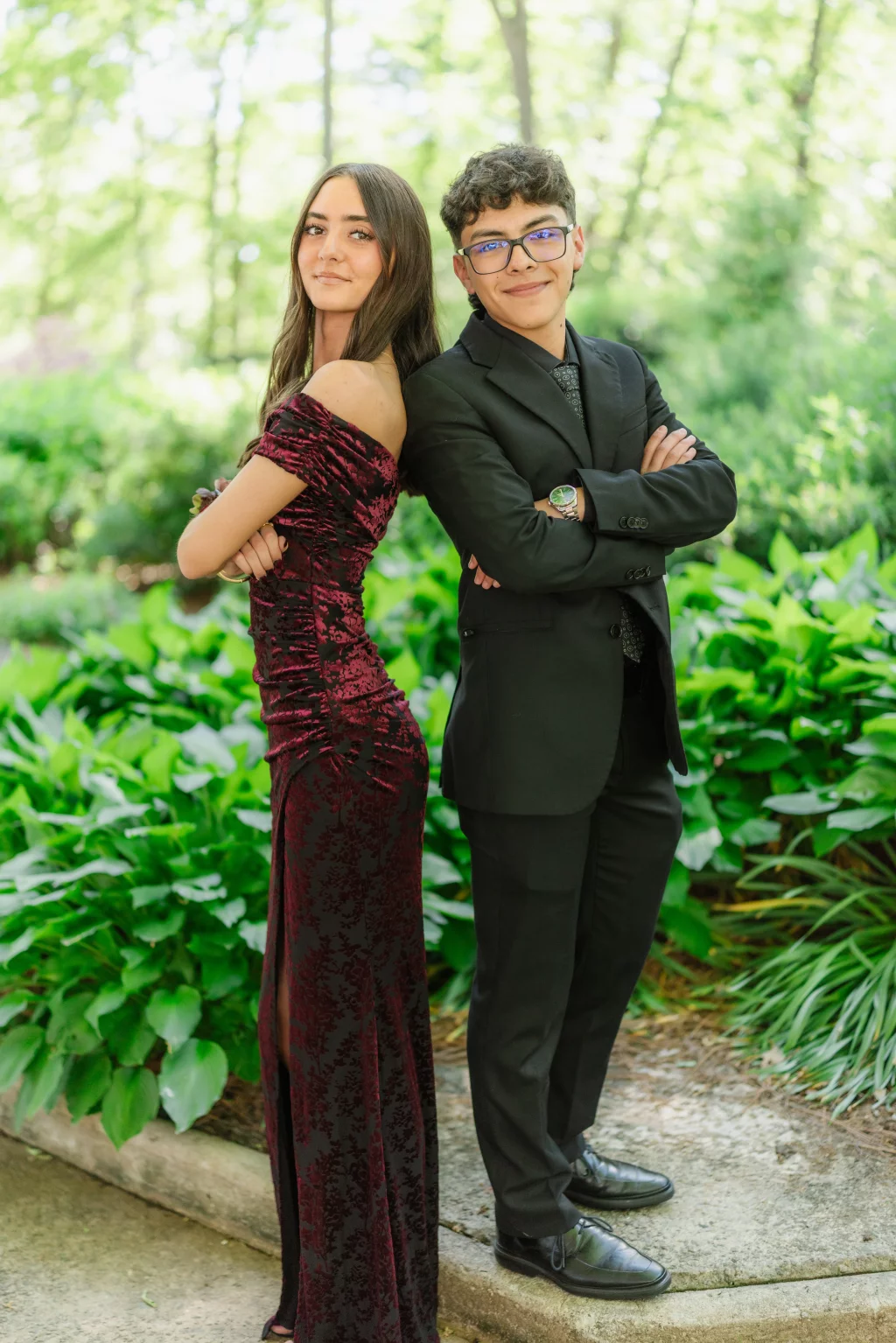 BJA Jr/Sr Banquet Prom at Sue Simpson Park: Two teenagers stand back to back with arms crossed, dressed formally; the girl wears an off-shoulder burgundy gown and the boy wears a black suit with glasses. They are outdoors with lush green plants in the background.
