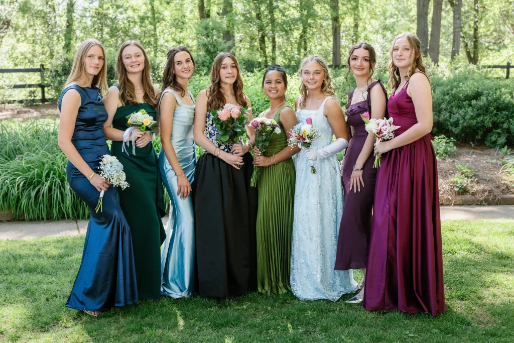 BJA Jr/Sr Banquet Prom at Sue Simpson Park: Eight young women in formal dresses stand together outdoors, each holding a bouquet of flowers, smiling at the camera with greenery and trees in the background.