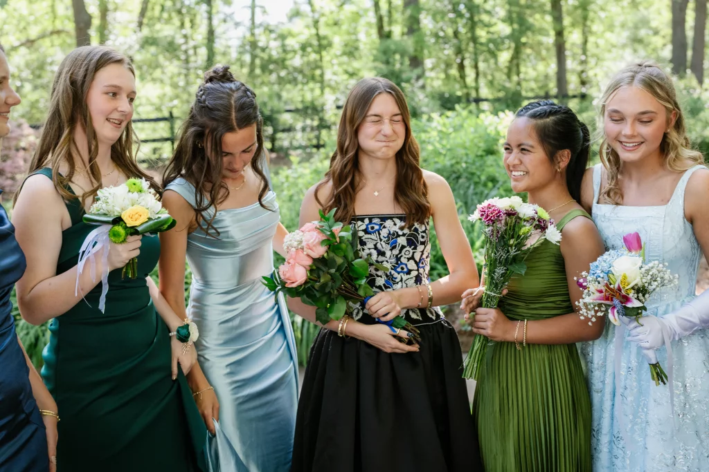 BJA Jr/Sr Banquet Prom at Sue Simpson Park: Five young women in colorful dresses stand outdoors holding bouquets, smiling and laughing together. Trees and greenery fill the background.