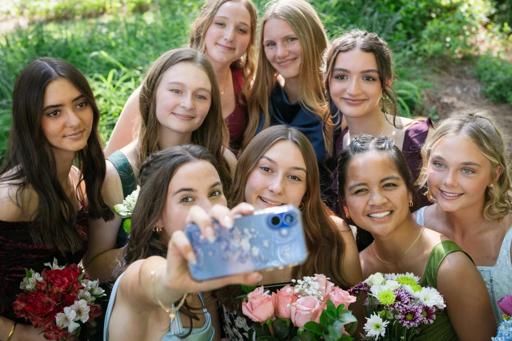 BJA Jr/Sr Banquet Prom at Sue Simpson Park: A group of nine young women dressed in formal attire gather closely together outdoors, smiling as one of them holds up a phone to take a selfie. Several are holding bouquets of flowers, with greenery visible in the background.
