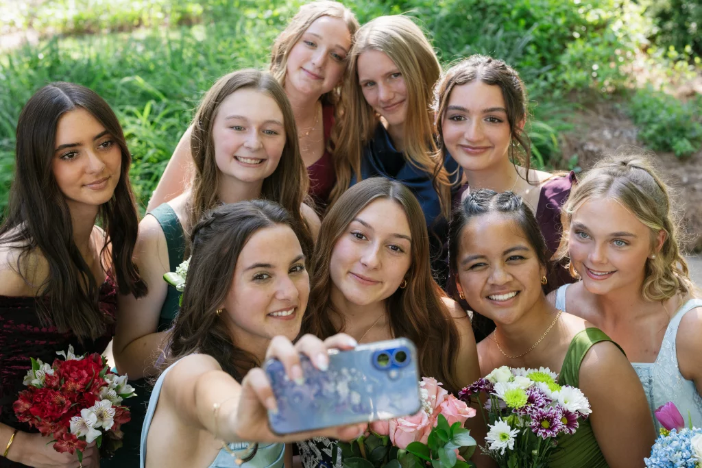 BJA Jr/Sr Banquet Prom at Sue Simpson Park: A group of nine young women in formal dresses, holding flower bouquets, smile together as one takes a selfie outdoors in a garden setting.