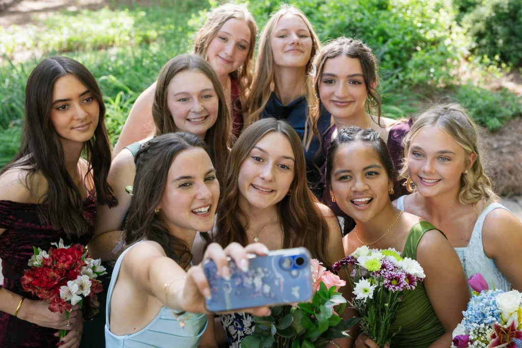 BJA Jr/Sr Banquet Prom at Sue Simpson Park: A group of nine teenage girls dressed in formal attire pose closely together outdoors, smiling as one takes a selfie. Several hold colorful bouquets of flowers, and greenery is visible in the background.