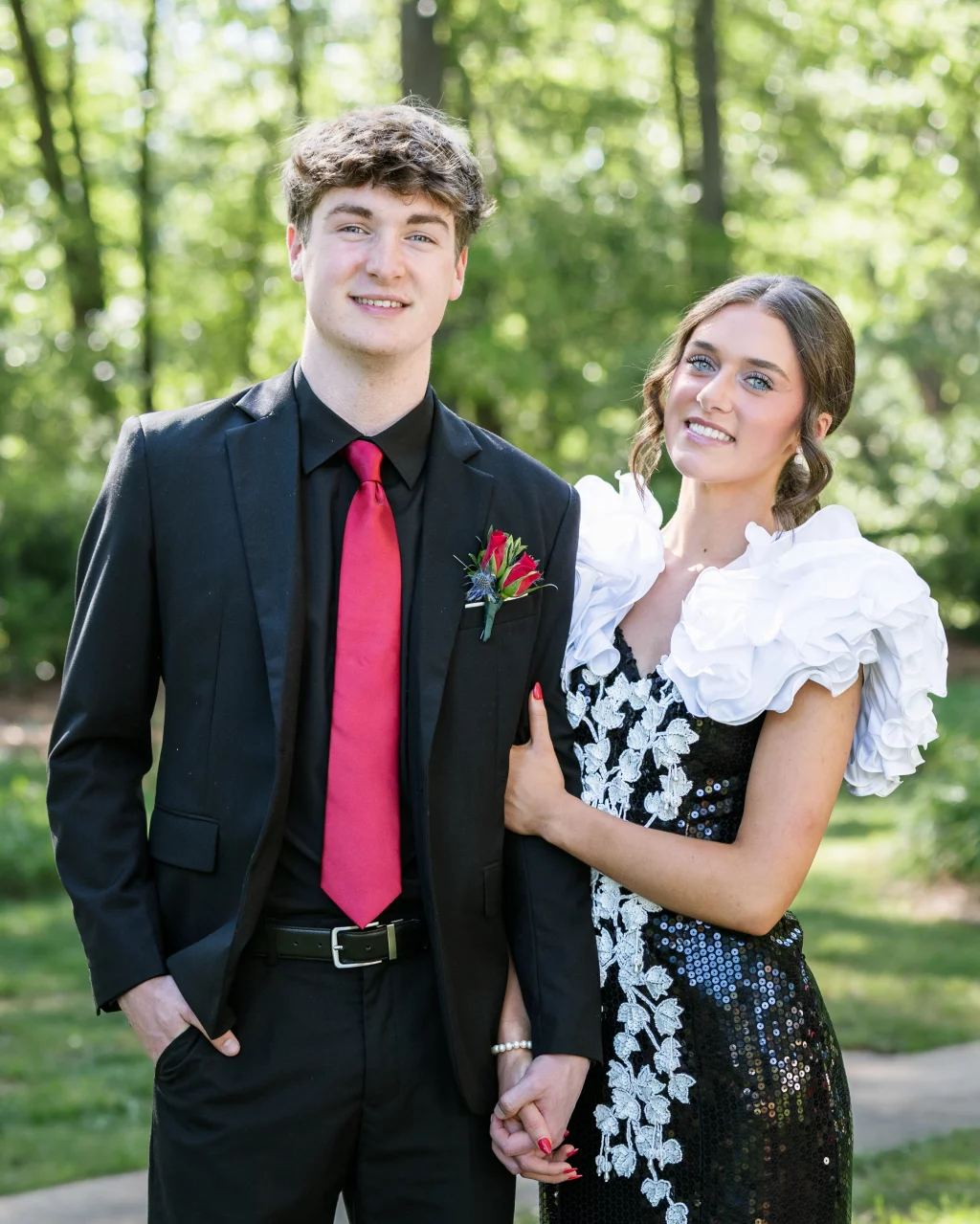 BJA Jr/Sr Banquet Prom at Sue Simpson Park: Two young people dressed formally, standing outdoors. The person on the left wears a black suit with a red tie and boutonniere, while the person on the right wears a black sequin dress with white floral accents and large ruffled white sleeves. They are holding hands and smiling at the camera. Trees and greenery are in the background.
