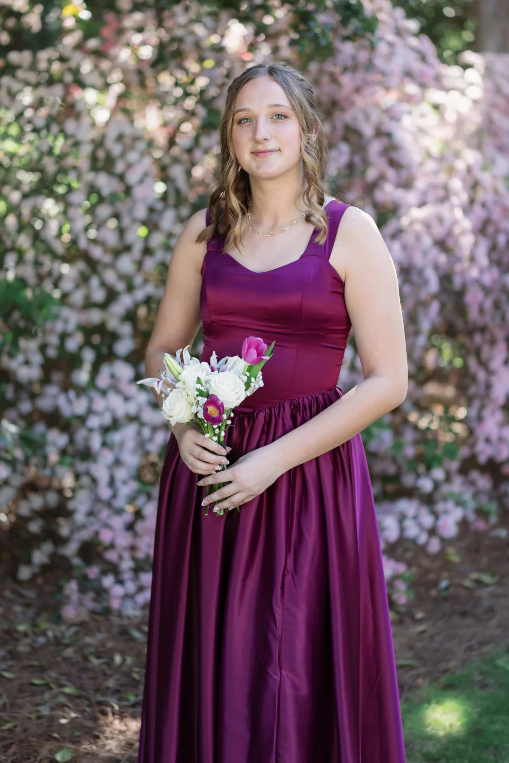 BJA Jr/Sr Banquet Prom at Sue Simpson Park: Young woman in a sleeveless magenta dress holding a bouquet of white and pink flowers, standing outdoors in front of blooming pink bushes.