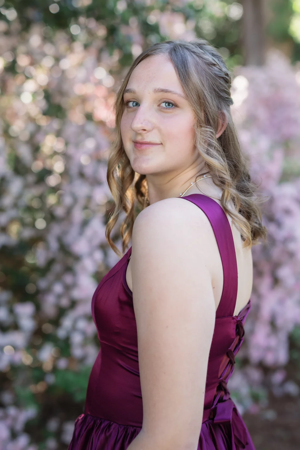 BJA Jr/Sr Banquet Prom at Sue Simpson Park: Young woman with wavy, light brown hair wearing a sleeveless, deep magenta dress, standing outdoors in front of soft-focus pink and green foliage, looking over her shoulder at the camera.