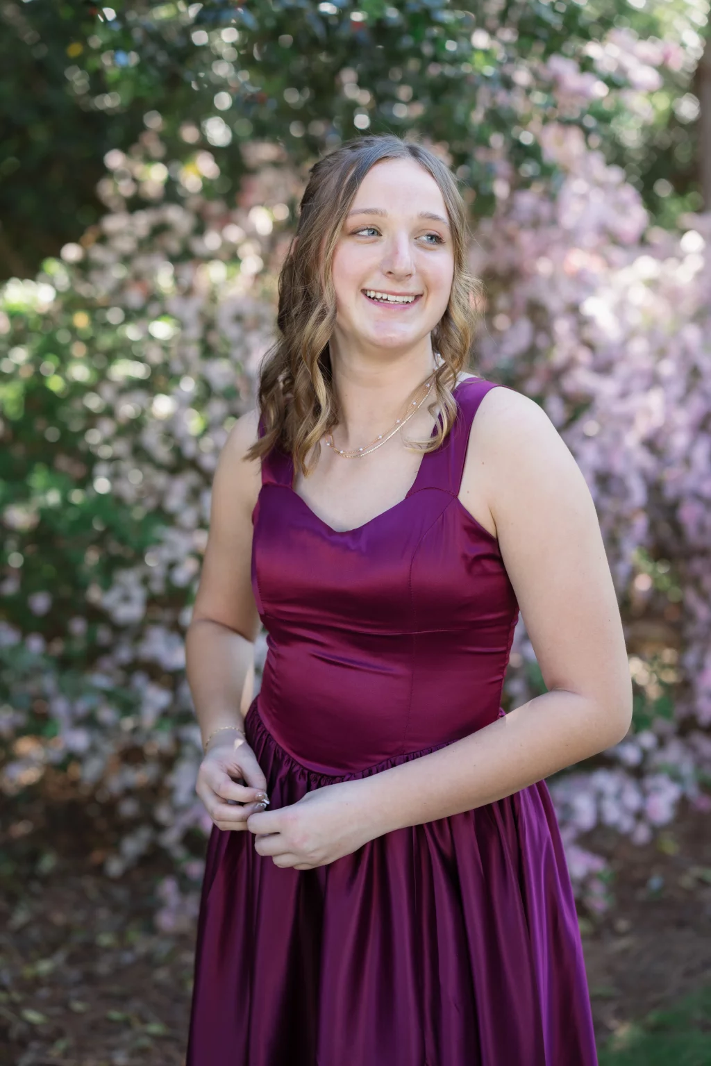 BJA Jr/Sr Banquet Prom at Sue Simpson Park: Young woman in a sleeveless, deep magenta dress standing outdoors in front of blooming pink flowers, smiling and looking slightly to the side.