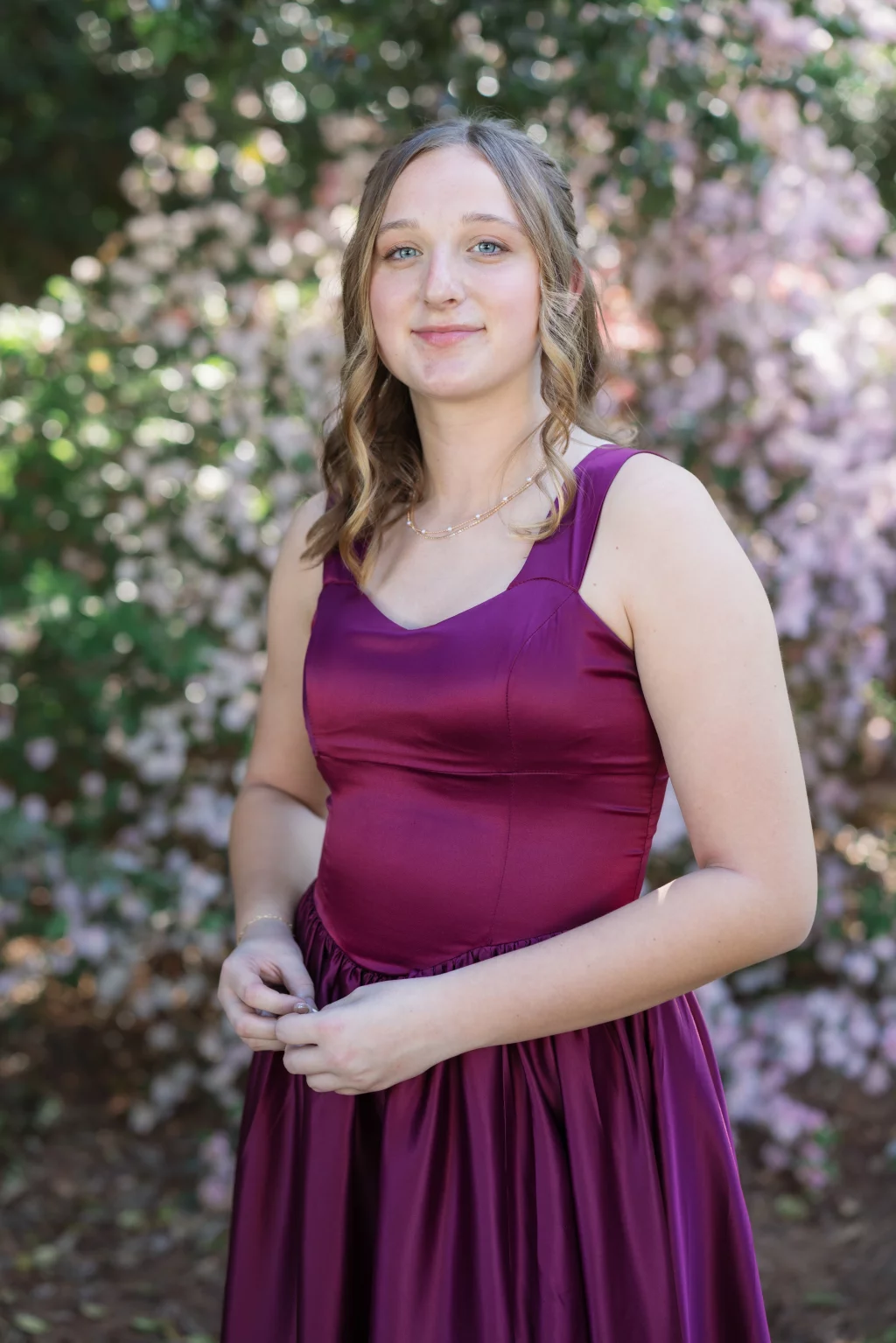 BJA Jr/Sr Banquet Prom at Sue Simpson Park: Young woman with light brown hair in loose curls, wearing a sleeveless, deep magenta dress, standing outdoors in front of blurred pink and green foliage.