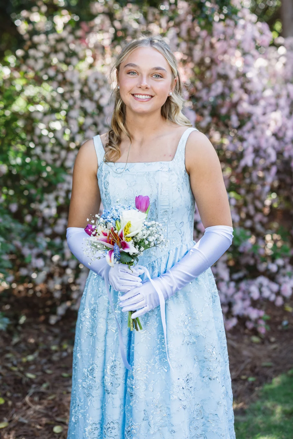 BJA Jr/Sr Banquet Prom at Sue Simpson Park: Young woman in a light blue formal dress and white gloves, holding a colorful bouquet, standing outdoors in front of blooming pink flowers.