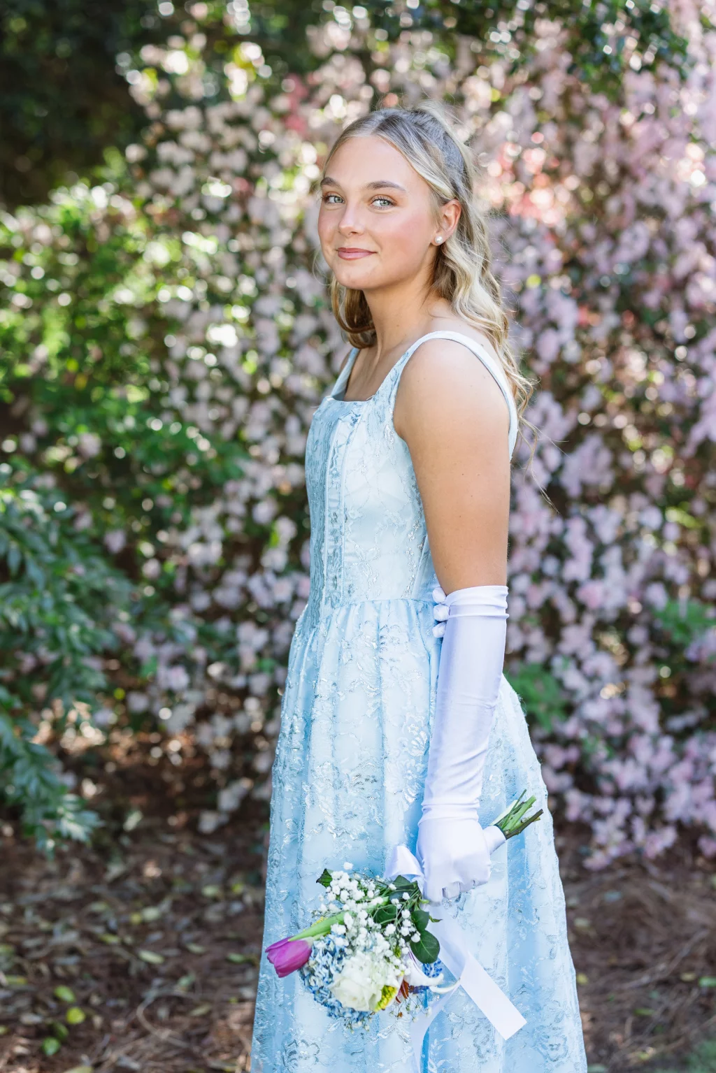 BJA Jr/Sr Banquet Prom at Sue Simpson Park: Young woman in a light blue lace dress and white gloves holding a small bouquet of flowers, standing outdoors in front of blooming pink and white bushes.