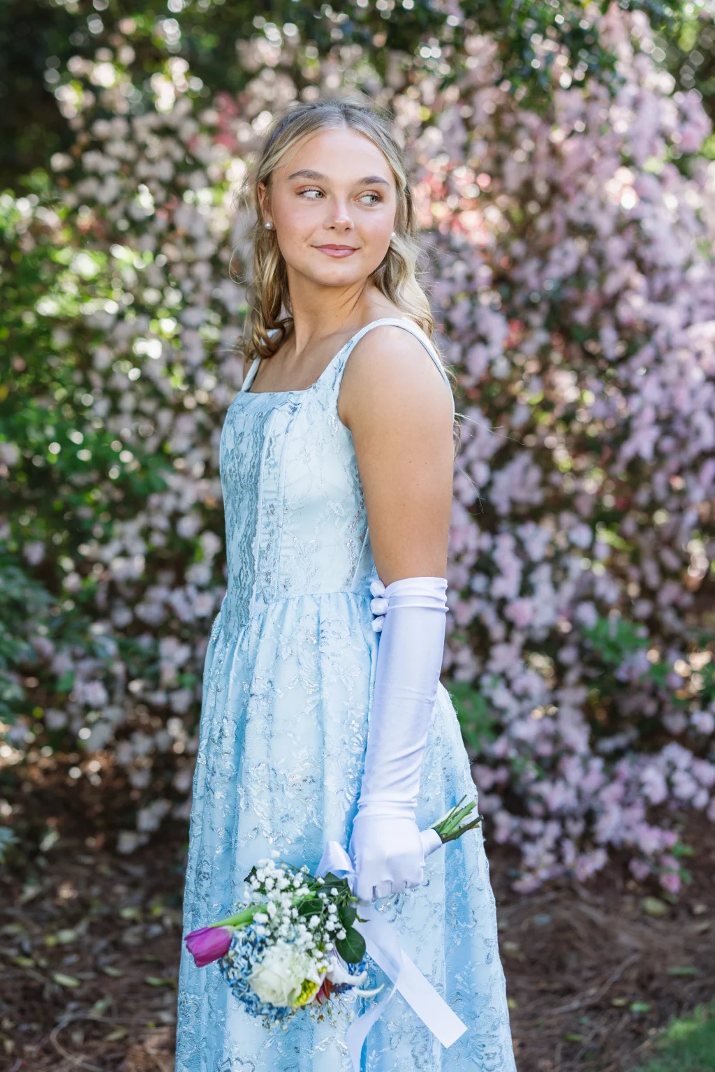 BJA Jr/Sr Banquet Prom at Sue Simpson Park: Young woman in a light blue lace dress and white gloves holding a small bouquet of flowers, standing outdoors in front of blooming pink bushes.