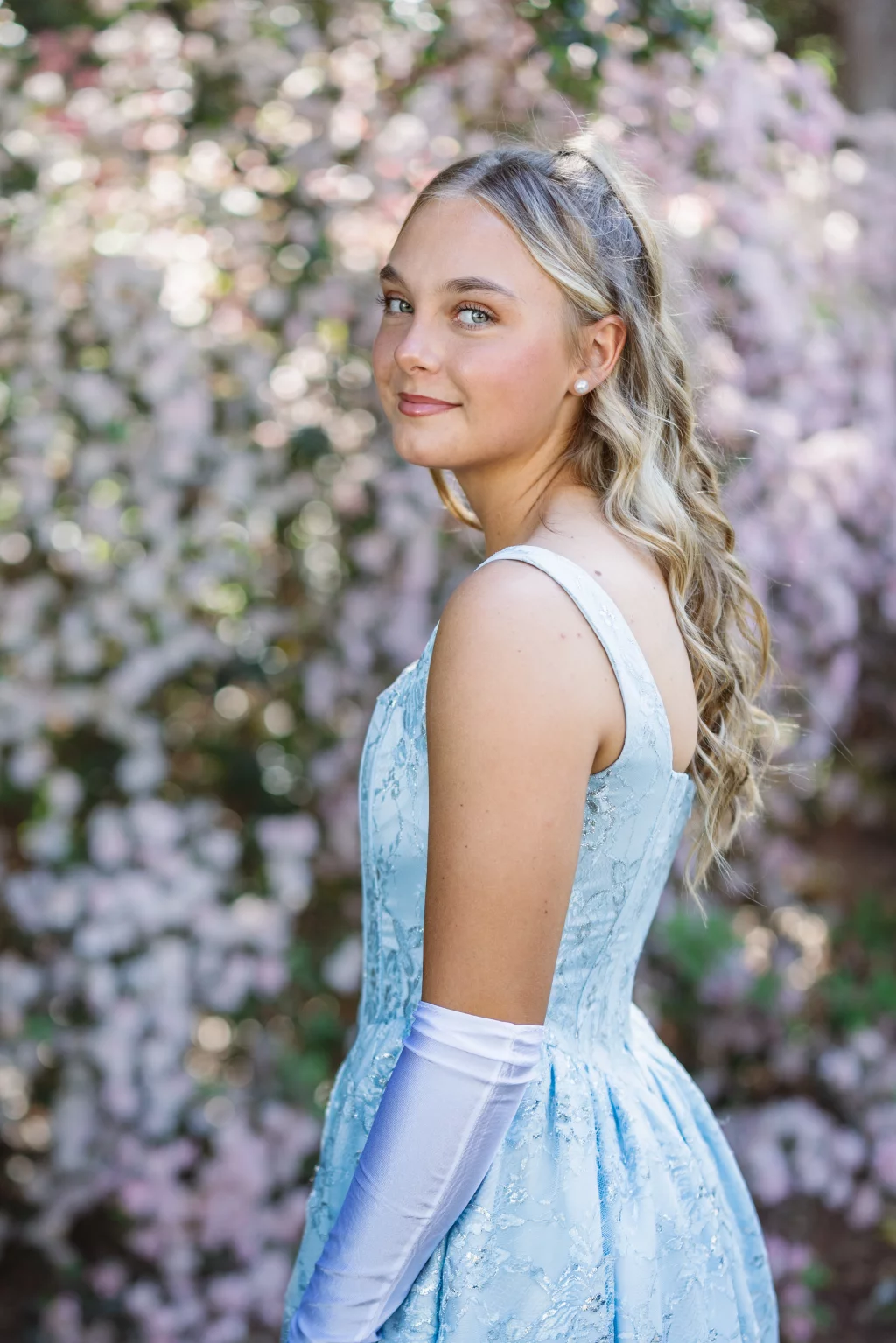BJA Jr/Sr Banquet Prom at Sue Simpson Park: Young woman with long blonde hair in a light blue formal dress and white gloves, standing outdoors in front of blooming pink flowers, looking over her shoulder at the camera.