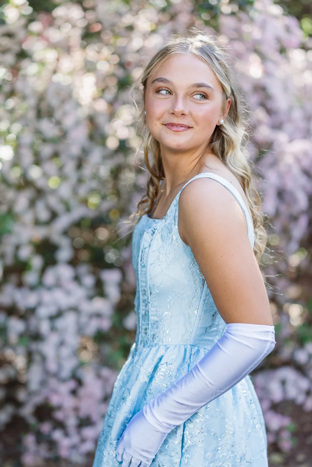 BJA Jr/Sr Banquet Prom at Sue Simpson Park: Young woman with long blond hair in a light blue formal dress and white opera gloves, standing outdoors in front of blooming pink flowers, looking over her shoulder and smiling.