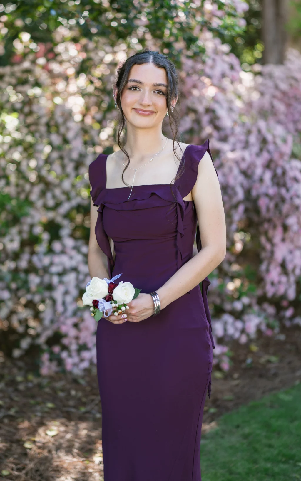 BJA Jr/Sr Banquet Prom at Sue Simpson Park: Young woman standing outdoors in front of pink flowering bushes, wearing a purple formal dress and holding a small bouquet of white and red flowers.