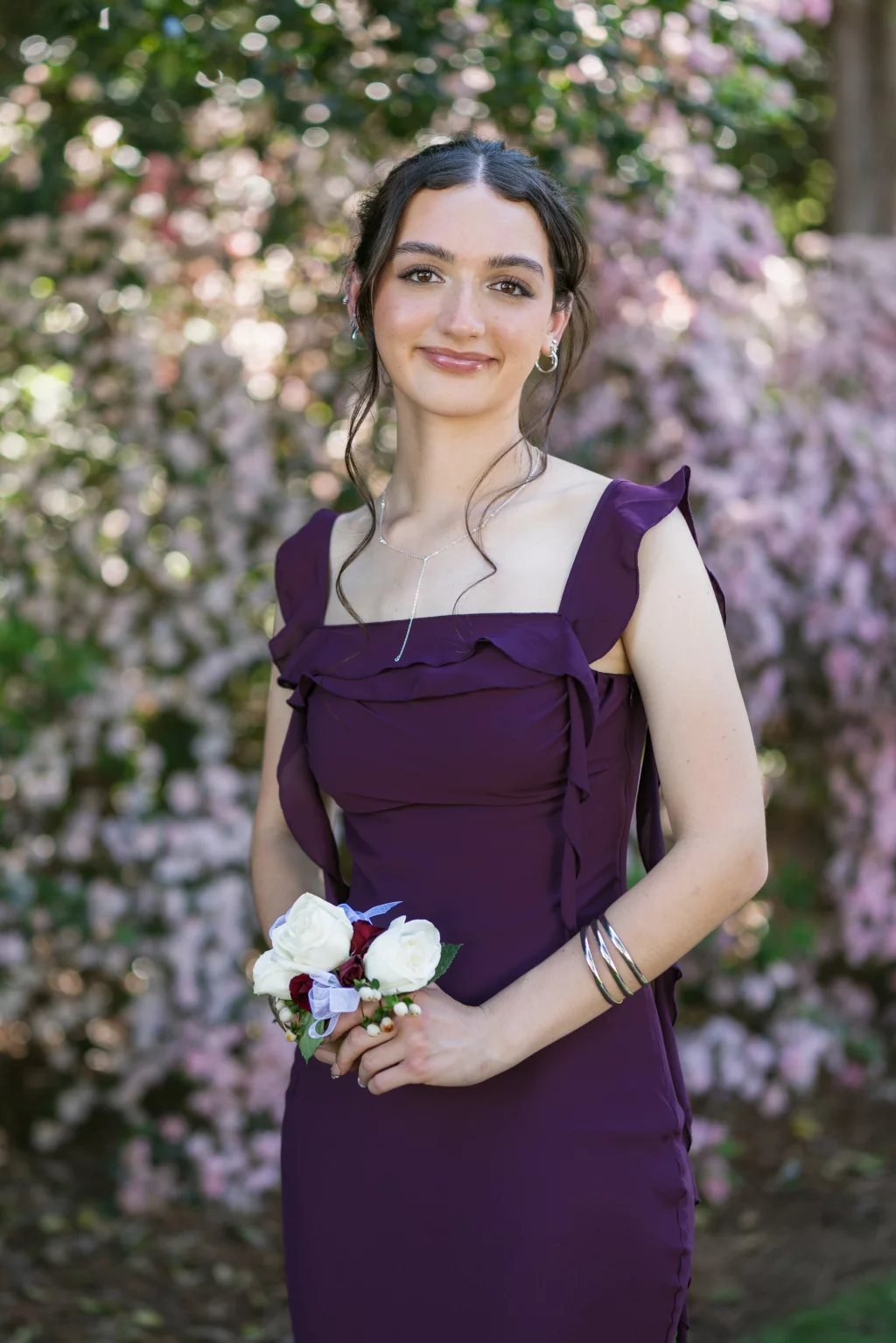 BJA Jr/Sr Banquet Prom at Sue Simpson Park: Young woman in a purple dress standing outdoors in front of pink and green foliage, holding a white and red flower corsage, smiling at the camera.