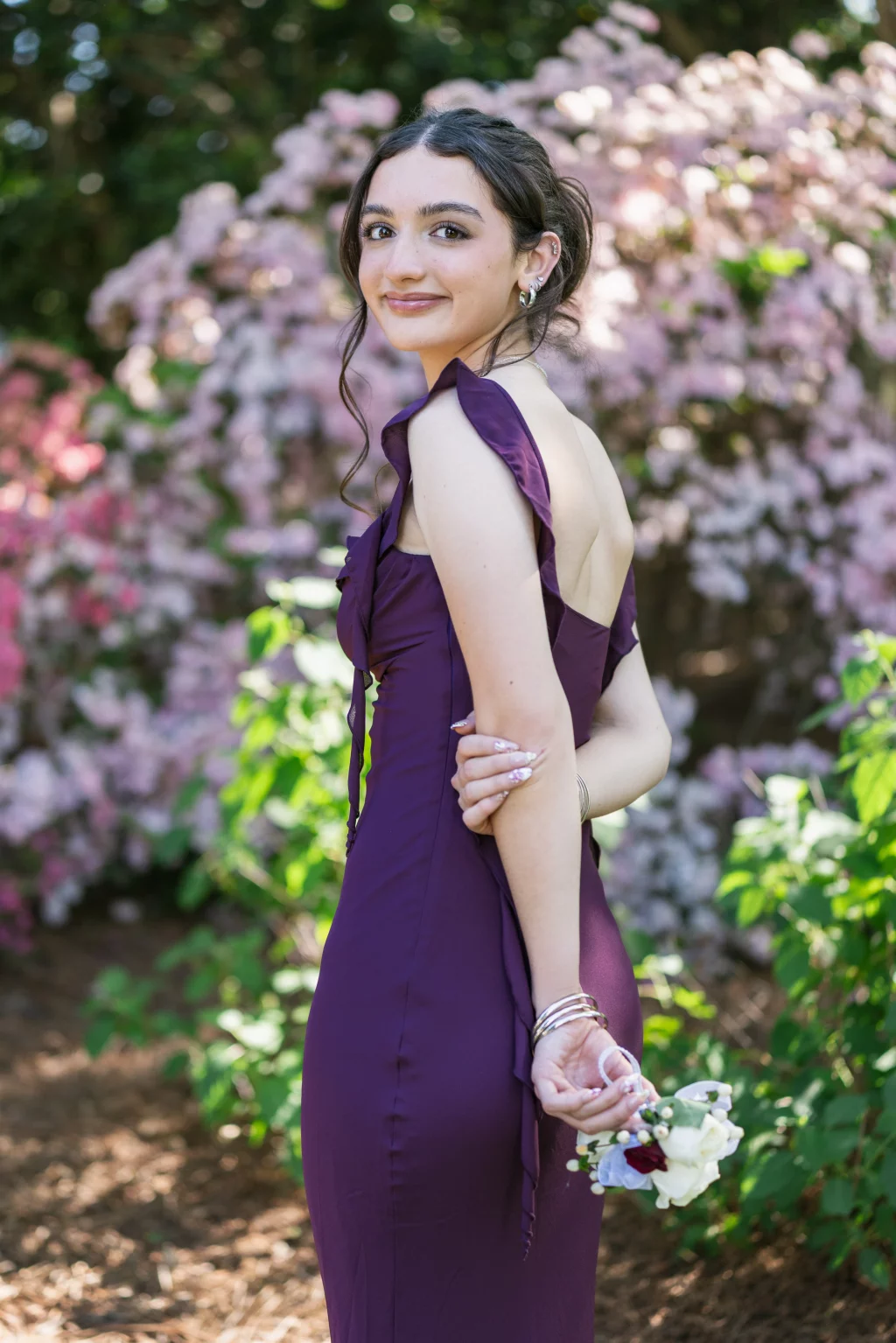 BJA Jr/Sr Banquet Prom at Sue Simpson Park: Young woman in a sleeveless purple dress standing outdoors, smiling over her shoulder, holding a small white and burgundy floral corsage, with pink and green flowering bushes in the background.