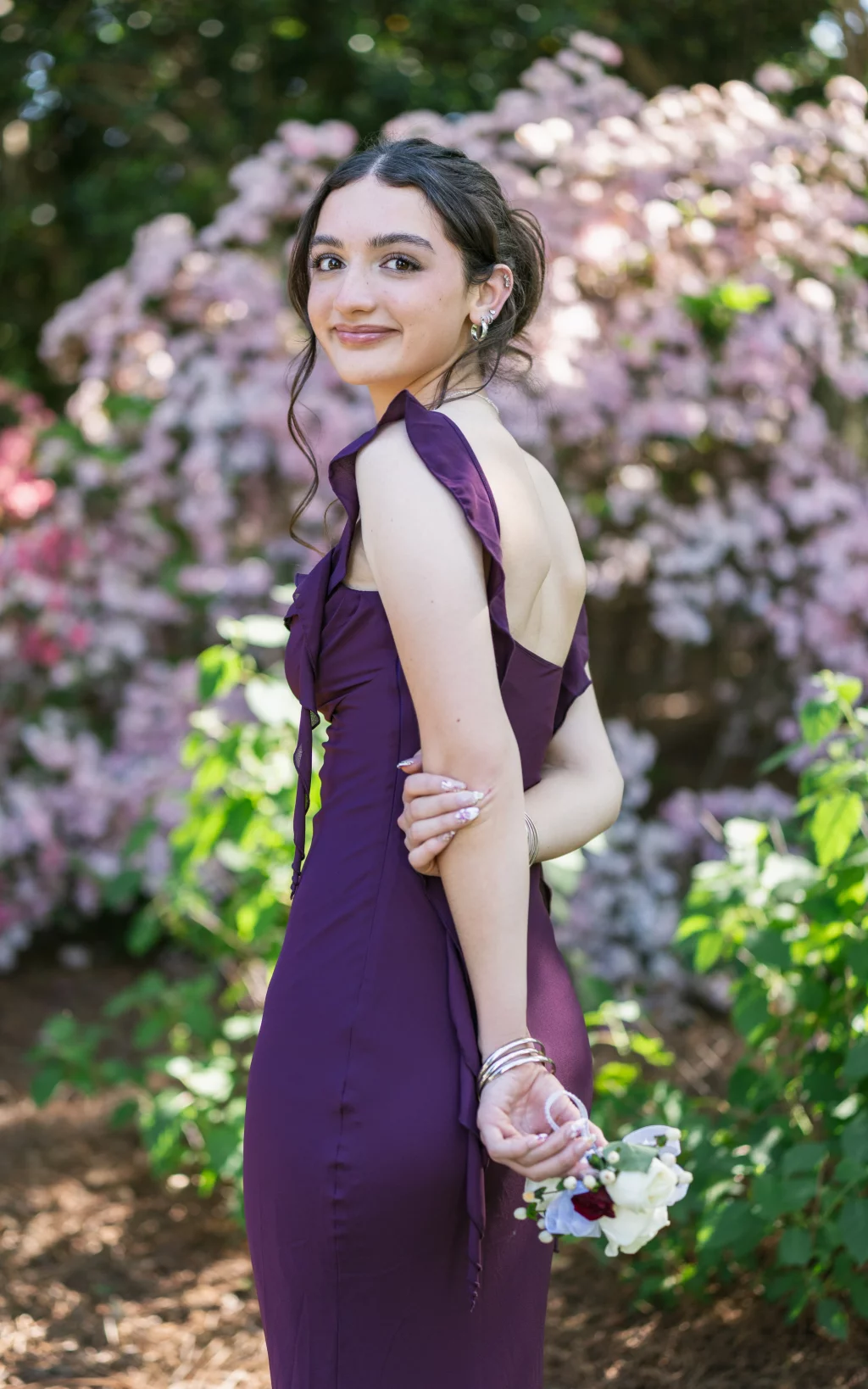 BJA Jr/Sr Banquet Prom at Sue Simpson Park: Young woman in a sleeveless purple dress standing outdoors, smiling over her shoulder, with a corsage in her hand and pink flowers in the background.