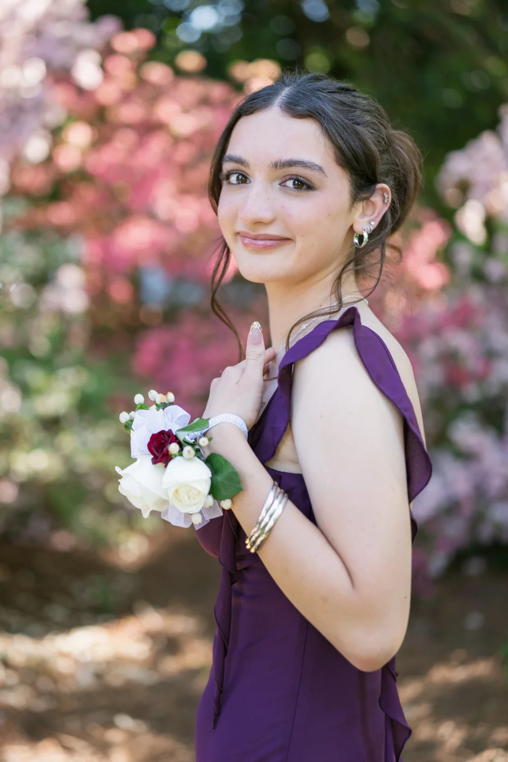 BJA Jr/Sr Banquet Prom at Sue Simpson Park: Young woman in a purple dress smiling at the camera, wearing a corsage with white and red flowers on her wrist, standing outdoors with pink and green blurred foliage in the background.