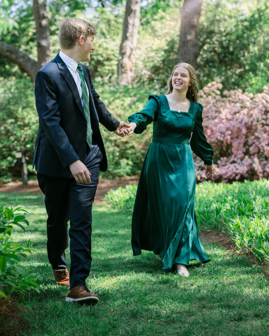 BJA Jr/Sr Banquet Prom at Sue Simpson Park: A young man in a dark suit and a young woman in a long green dress hold hands and smile while walking together in a sunlit garden.