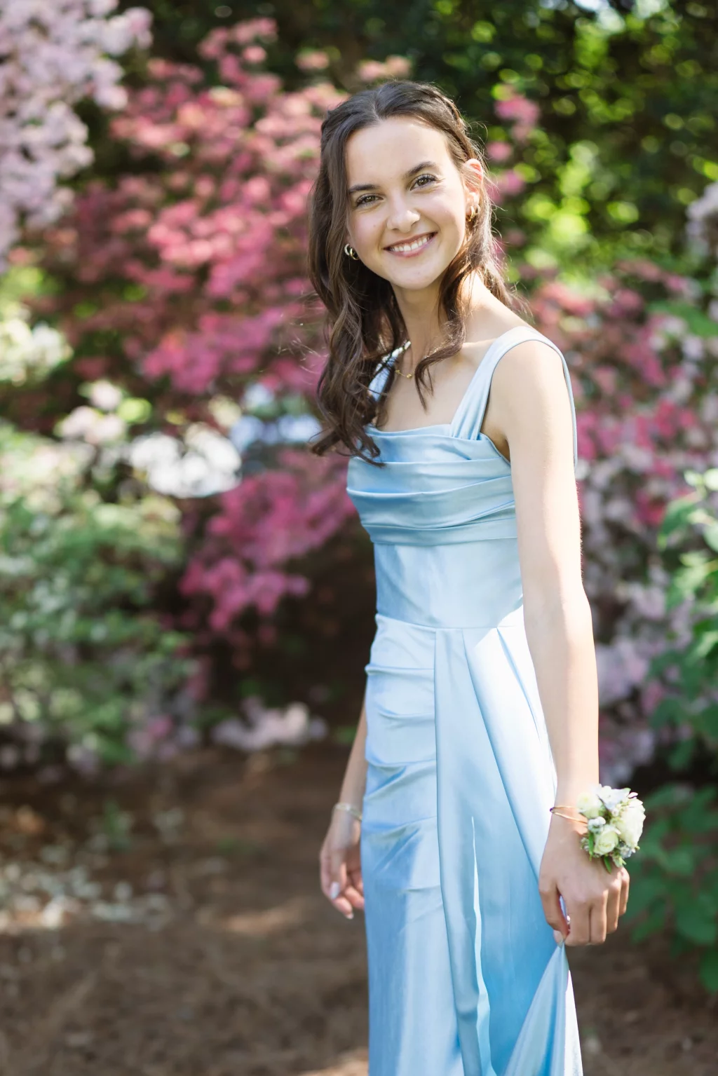 BJA Jr/Sr Banquet Prom at Sue Simpson Park: Young woman in a light blue formal dress, wearing a floral corsage, smiling outdoors with pink and green flowering bushes in the background.