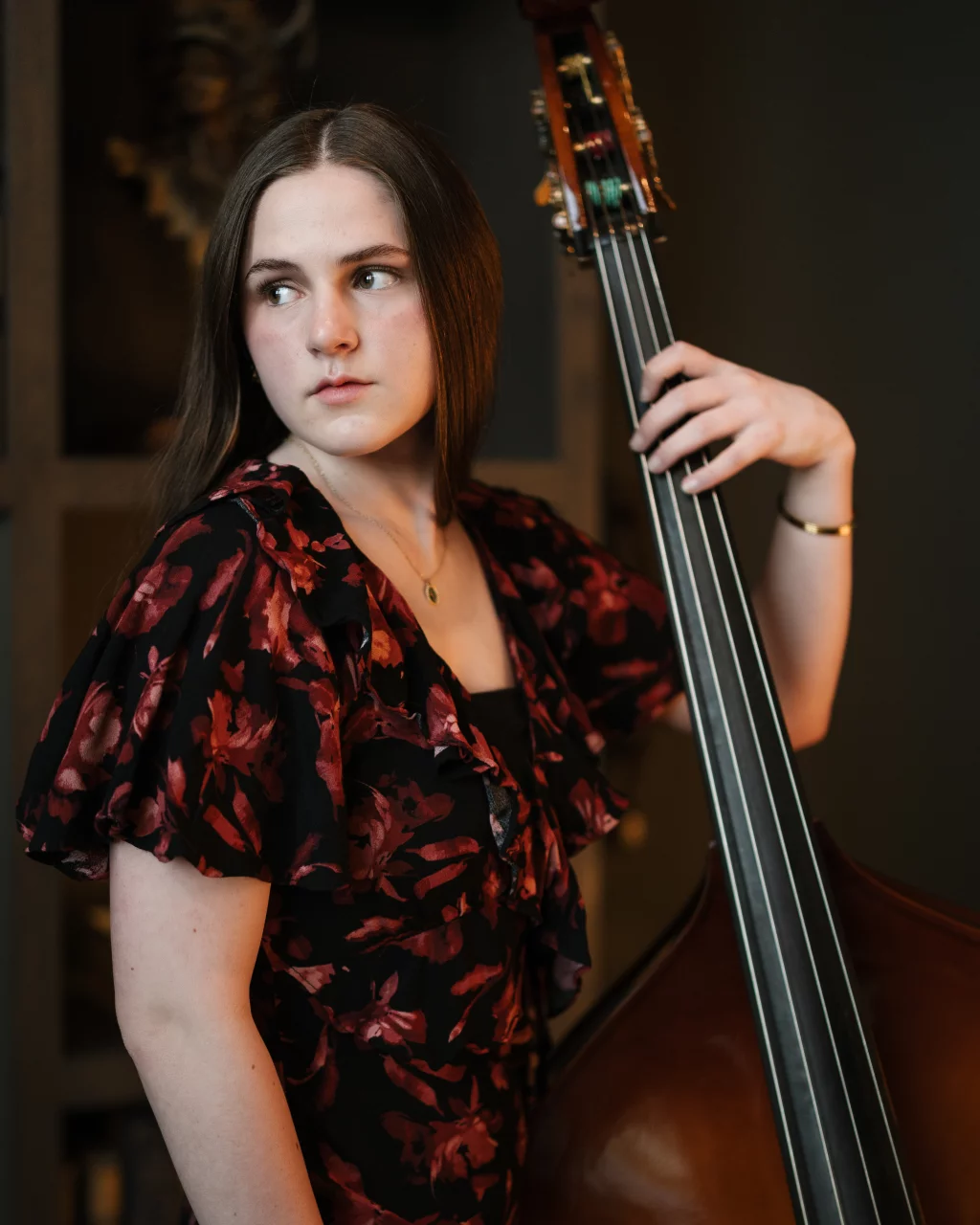 Kellis's senior photos taken Downtown, Greenville, SC: Young woman with straight brown hair wearing a black and red floral dress, holding an upright bass and looking to her left.