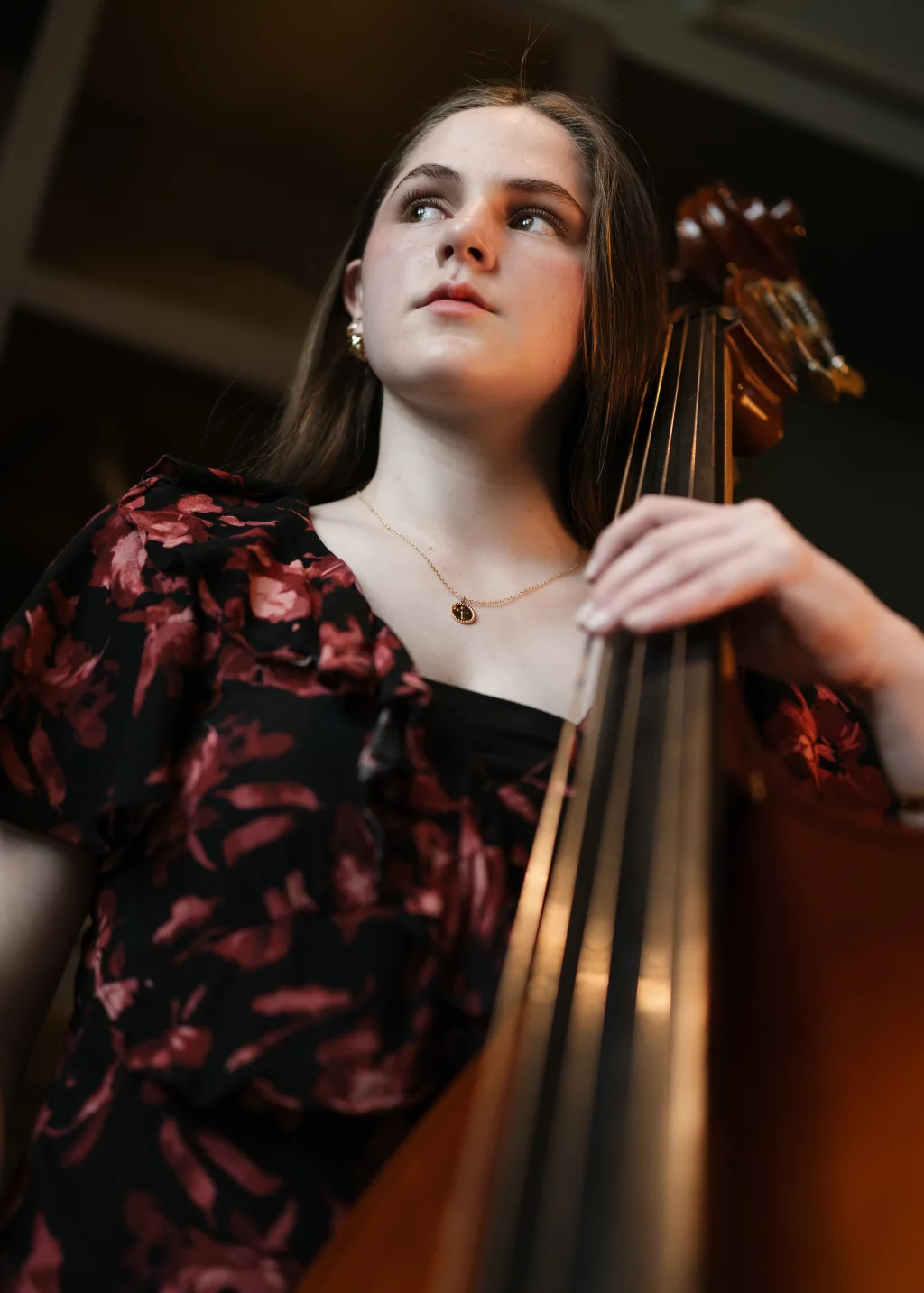 Kellis's senior photos taken Downtown, Greenville, SC: A young woman wearing a black and red floral blouse holds an upright bass, looking off to the side with a thoughtful expression.