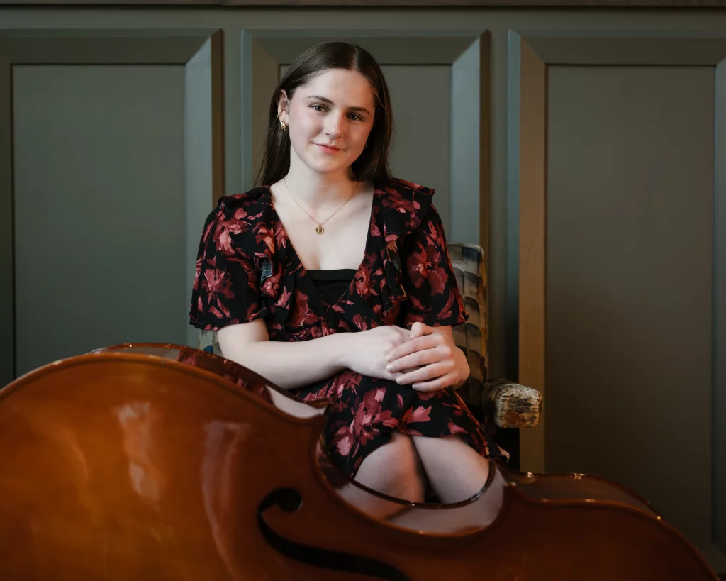 Kellis's senior photos taken Downtown, Greenville, SC: Young woman with long brown hair in a floral dress sits with her hands folded, a cello in the foreground.