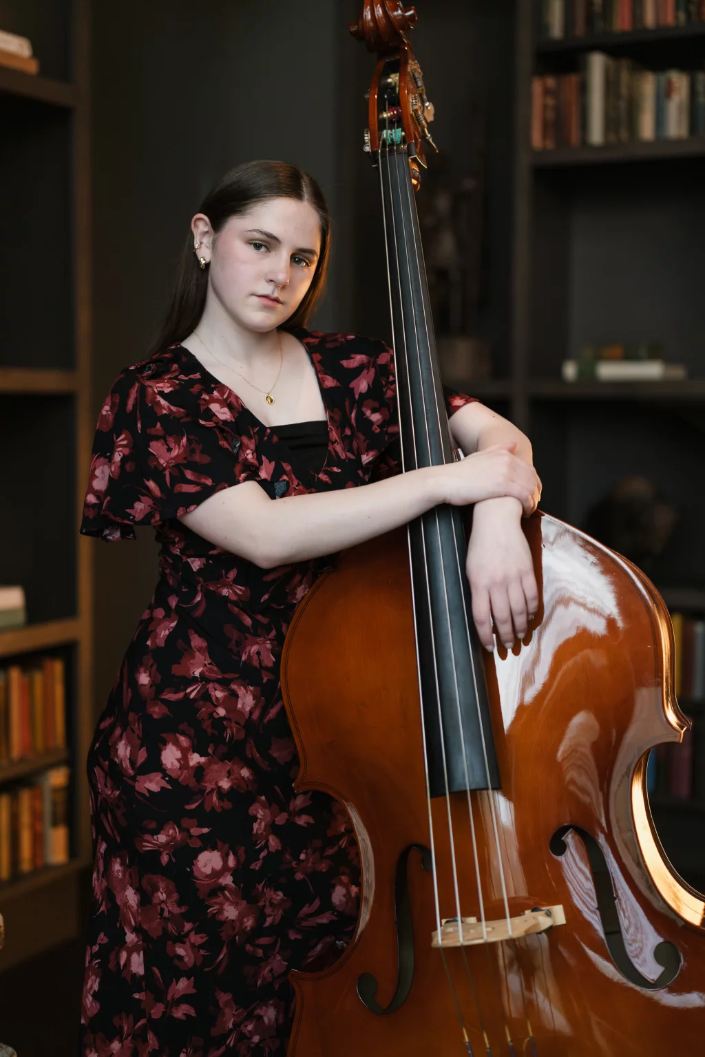 Kellis's senior photos taken Downtown, Greenville, SC: Young woman in a floral dress stands indoors, holding an upright double bass, with bookshelves in the background.
