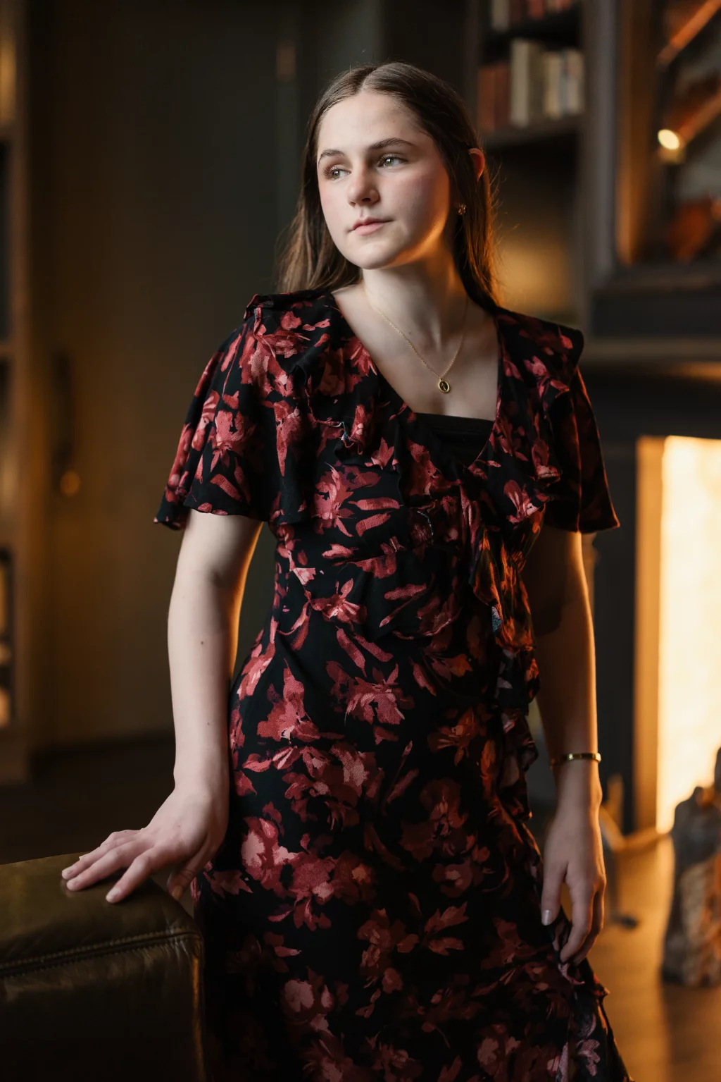Kellis's senior photos taken Downtown, Greenville, SC: Young woman with long brown hair wearing a black dress with red floral patterns, standing indoors near a fireplace and bookshelves, looking thoughtfully to the side.