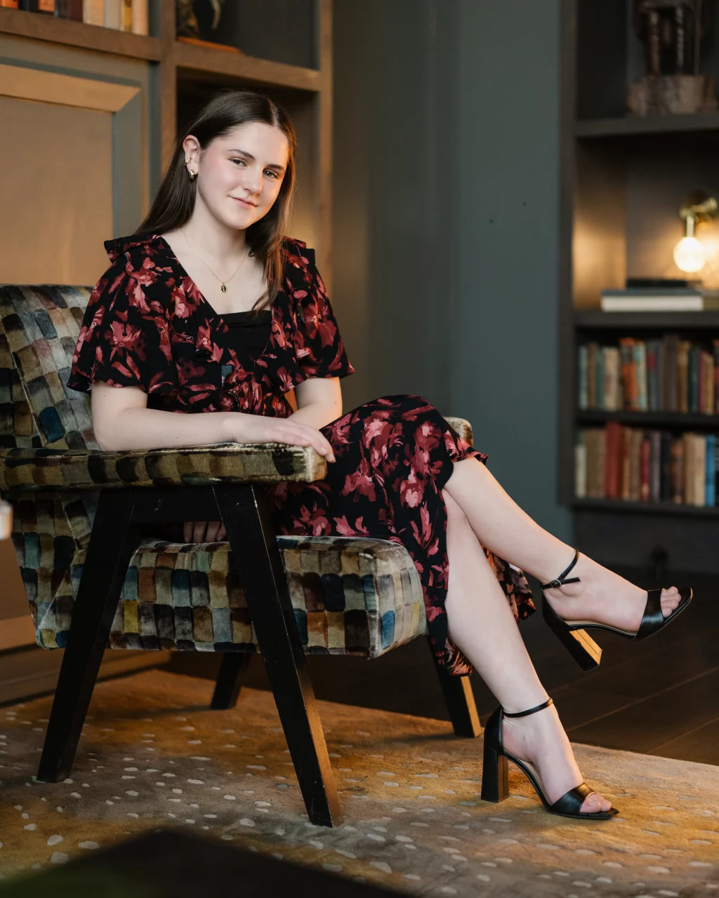 Kellis's senior photos taken Downtown, Greenville, SC: Young woman with straight brown hair wearing a floral dress and black heels, sitting sideways on a patterned armchair in a cozy room with bookshelves and soft lighting.