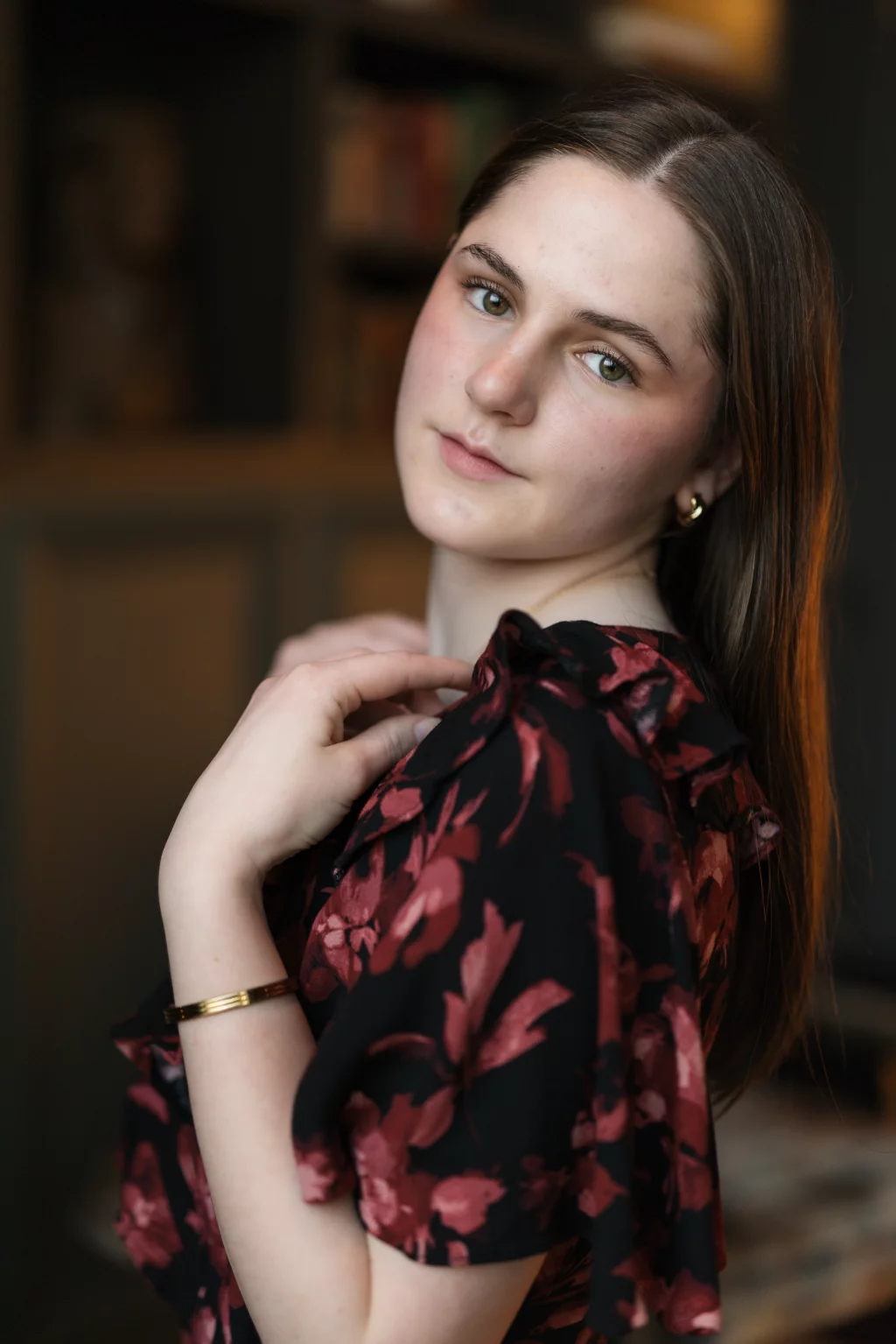 Kellis's senior photos taken Downtown, Greenville, SC: A young woman with long brown hair and light skin looks at the camera, wearing a black dress with a red floral pattern, gold earrings, and a gold bracelet, with a softly blurred background.